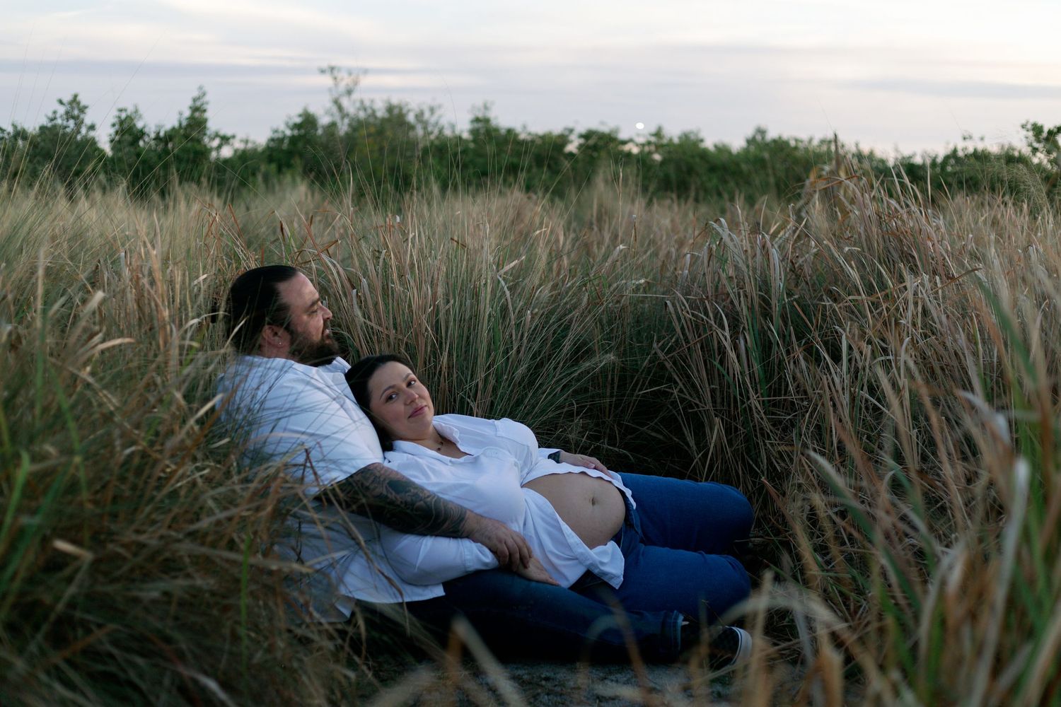 Two people relaxing together in tall grass meadow at dusk, lying peacefully among nature.