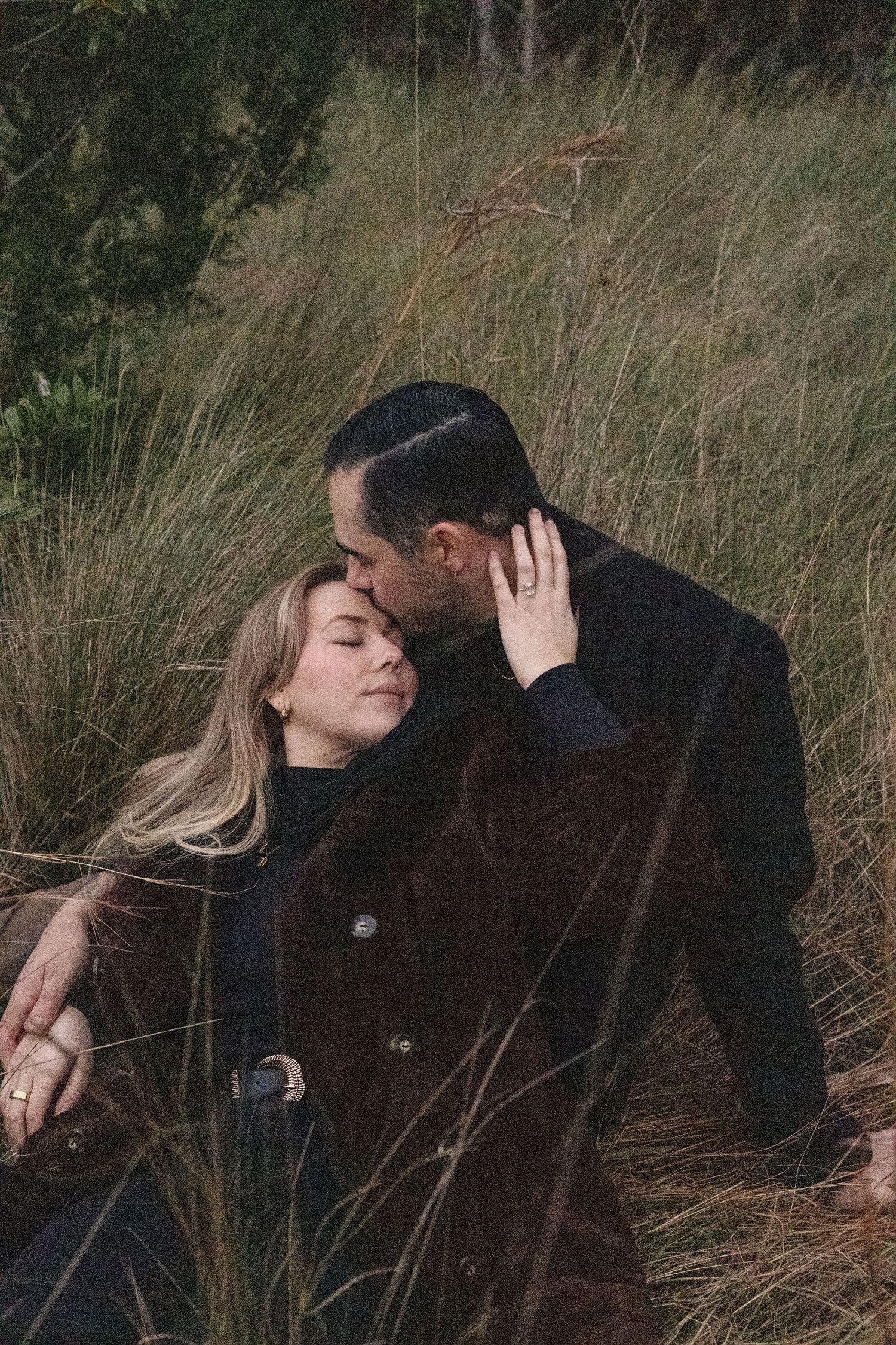 Couple sharing intimate moment in tall grass field during autumn wearing dark coats.