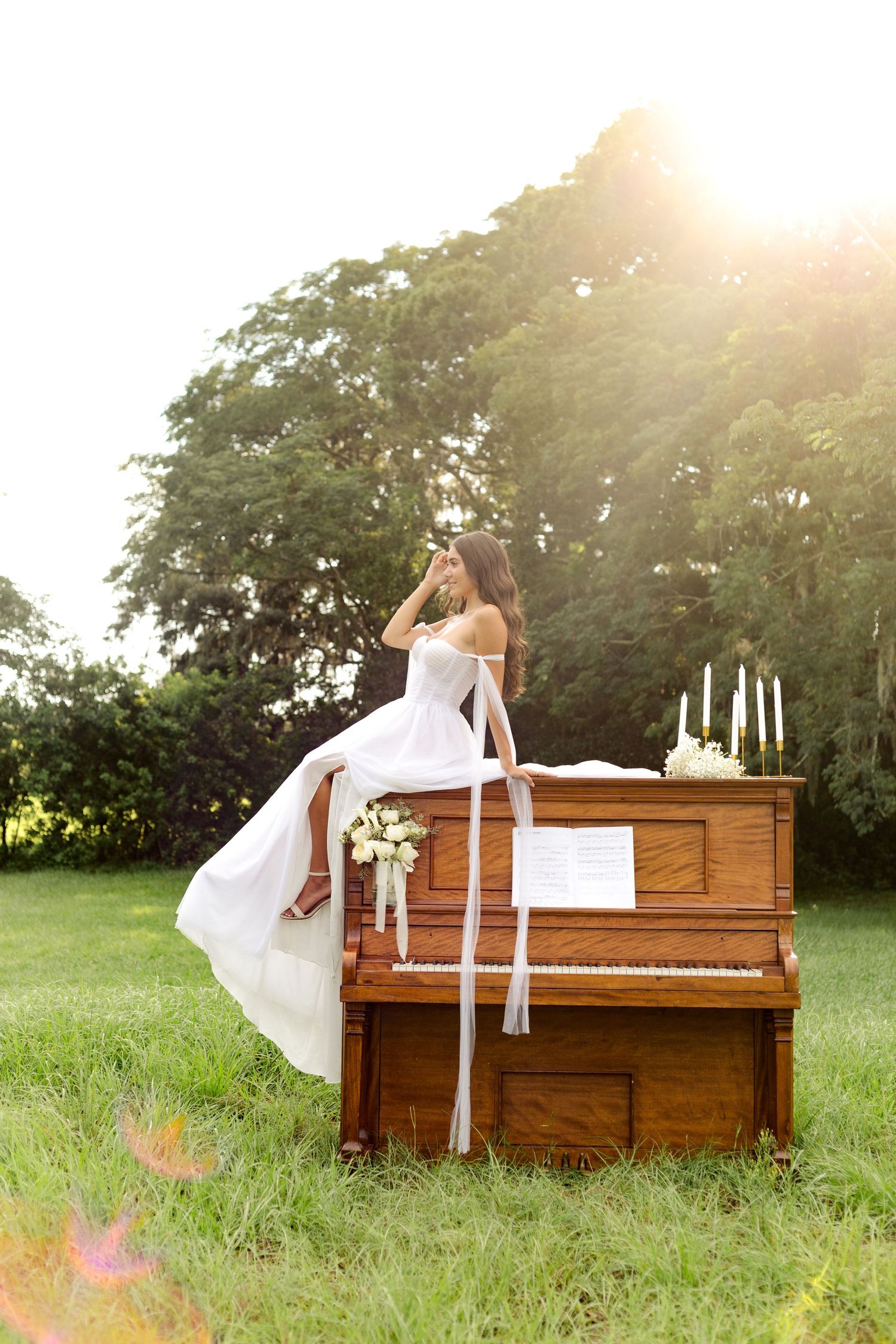 Vintage upright piano sits in grassy field with white candles and floral decoration under sunlit trees.