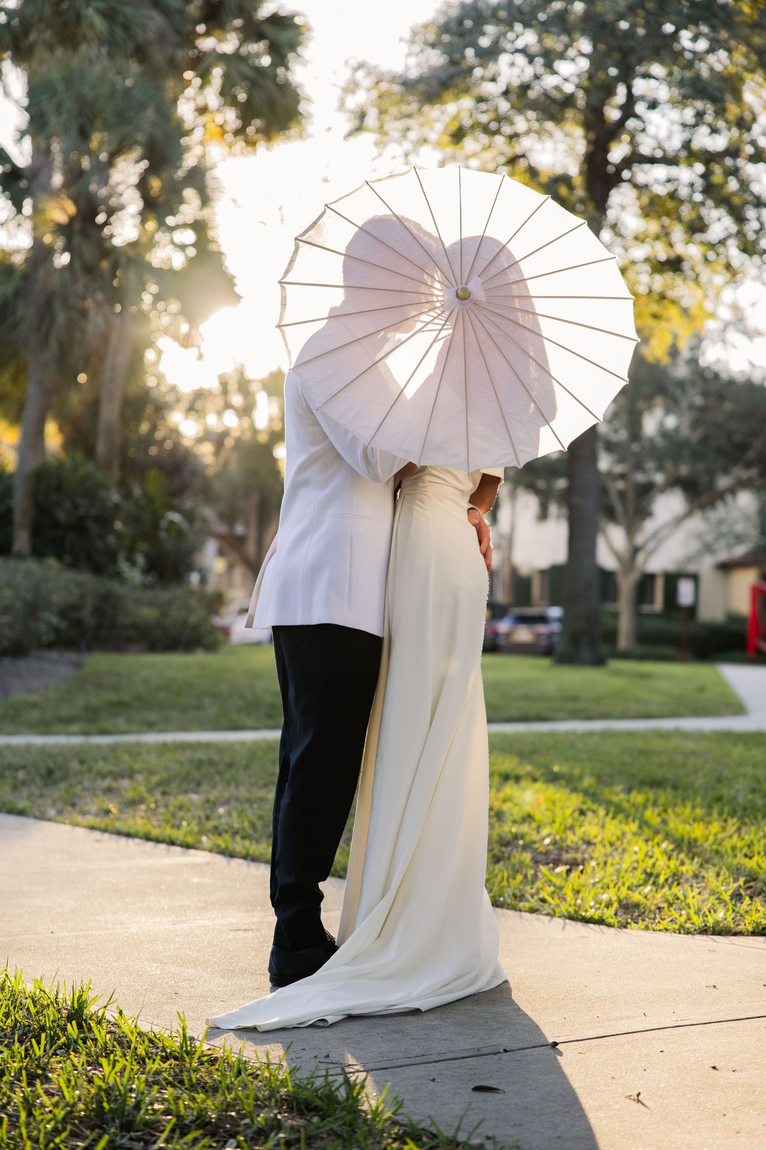 A couple shares a romantic moment under a white paper parasol during an outdoor wedding photo session at sunset.