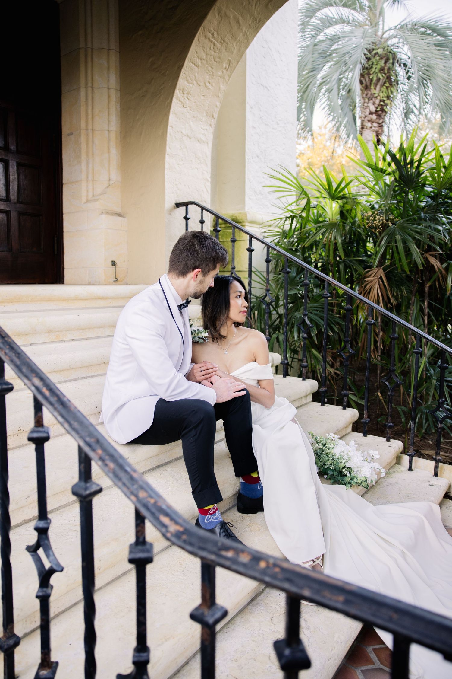 A couple in formal attire sits together on stone steps with an ornate iron railing and tropical plants in the background.