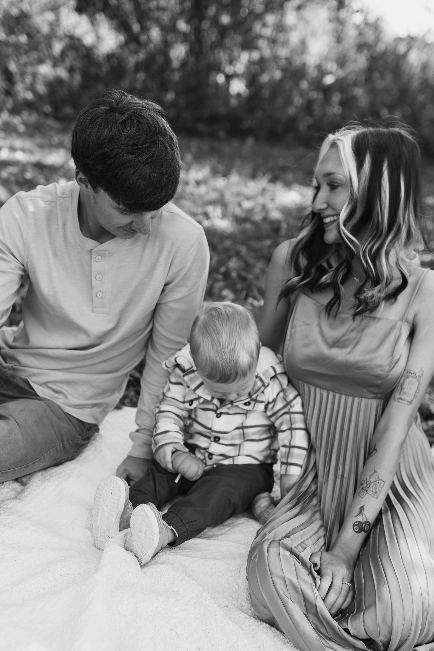 Black and white family photo of parents smiling at baby sitting on blanket outdoors.
