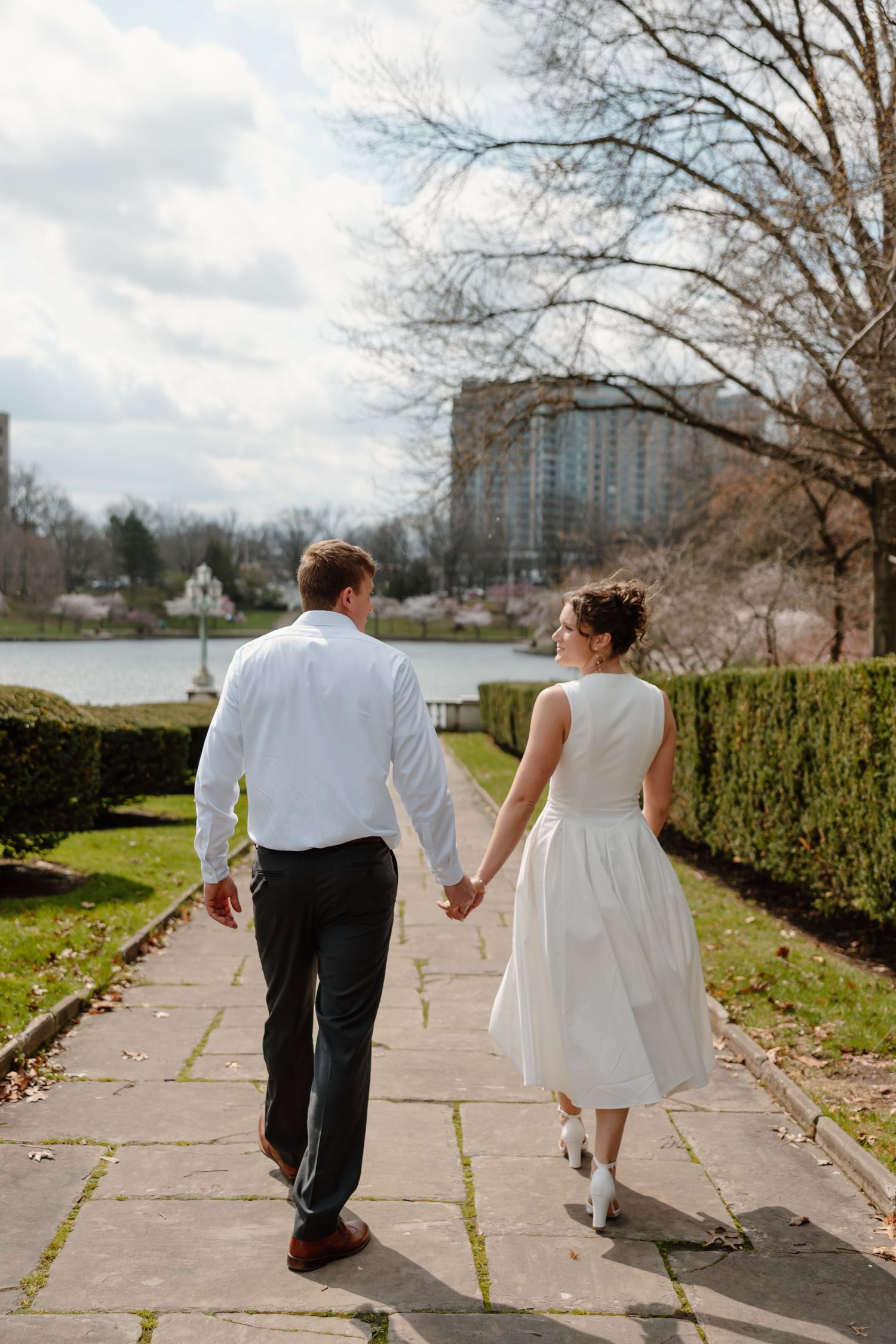 A couple in wedding attire holds hands walking along a garden path near a lake with city buildings in the background.
