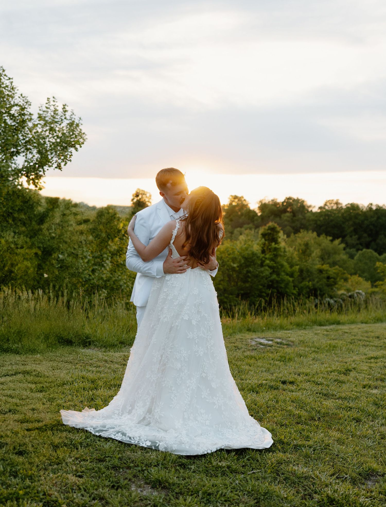Bride and groom embrace at golden hour sunset in lush green field, bride wearing elegant lace wedding gown with train.