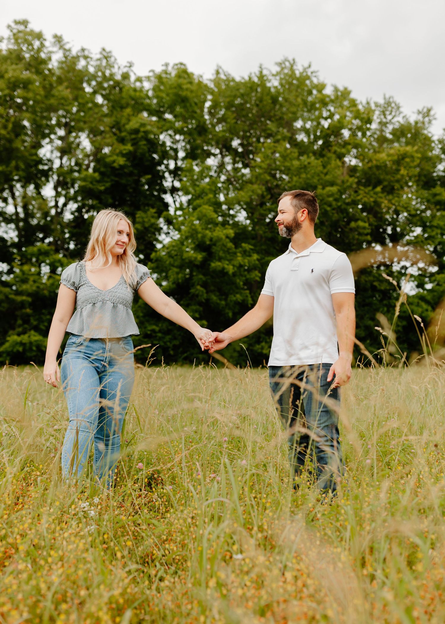 Casual couple in jeans holds hands while standing in sunlit meadow.