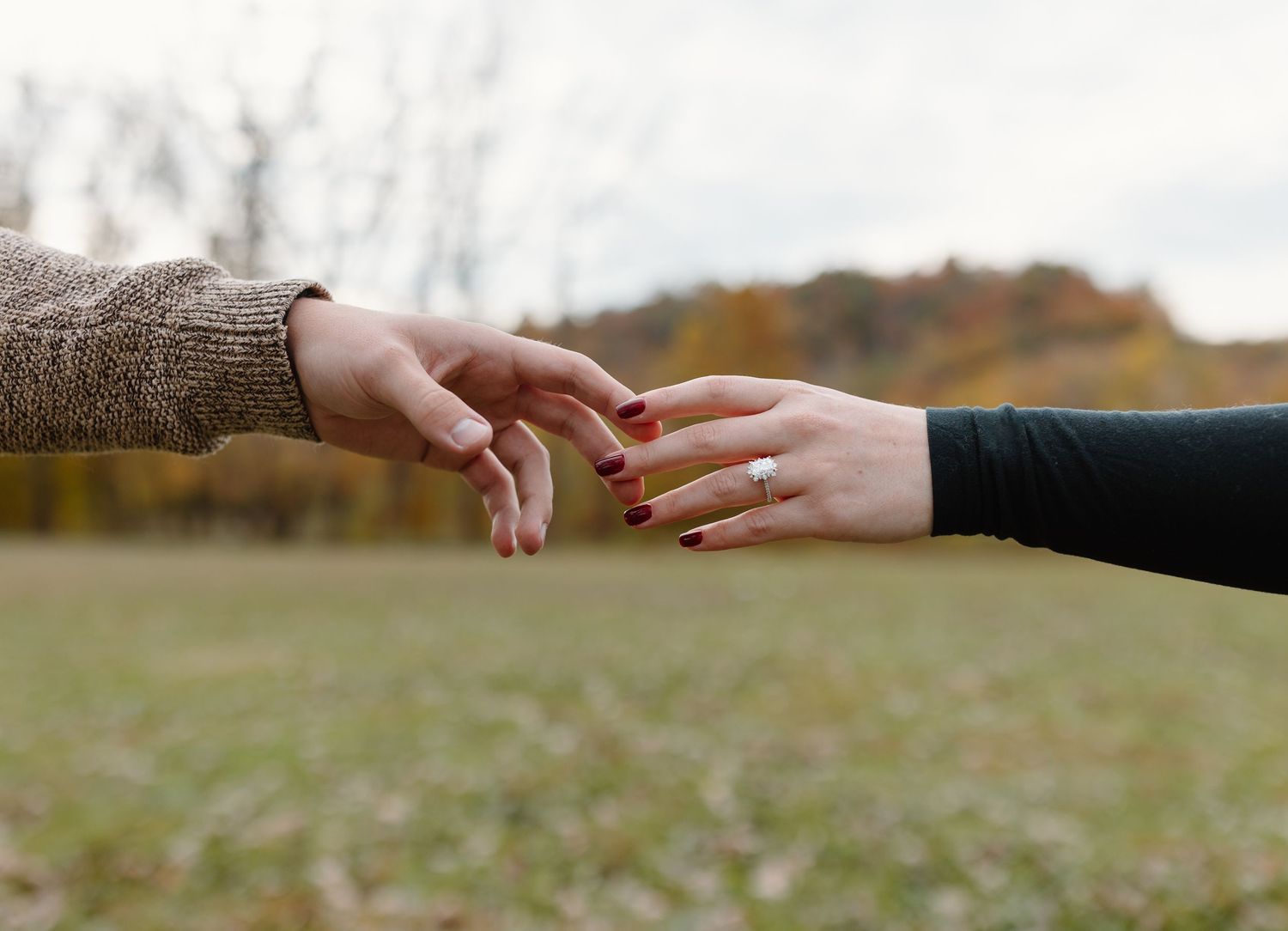 Two people with red nails gently touching fingertips outdoors in an autumn setting with colorful trees in background.