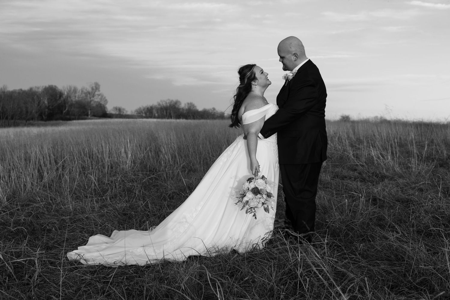 Black and white wedding photo of bride and groom embracing in a field at dusk, bride holding bouquet.