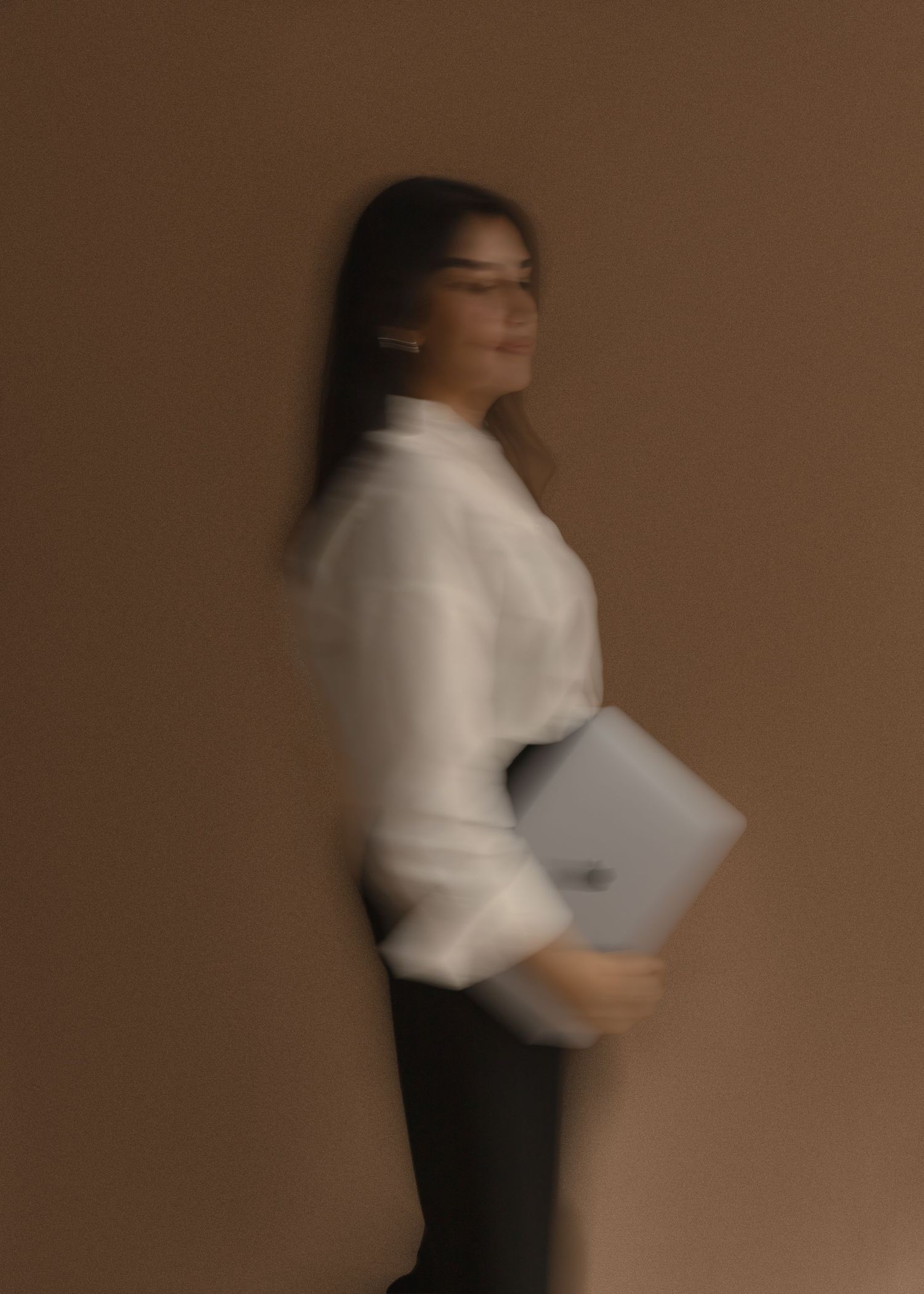 A professional woman in a white blouse holds a laptop, posing sideways against a warm brown background.