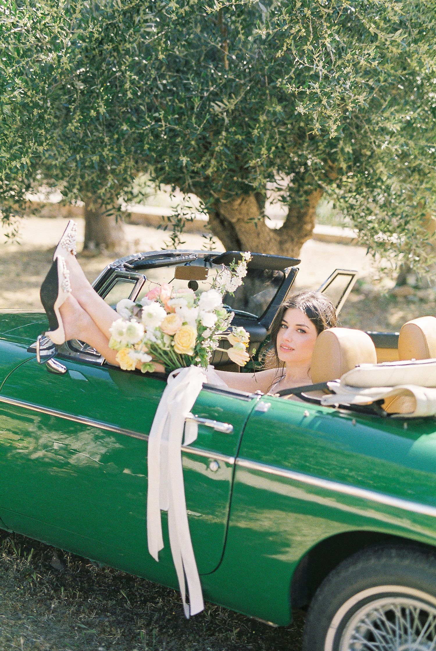 Bride lounging in a vintage green convertible car adorned with a floral bouquet and white ribbons under an olive tree.