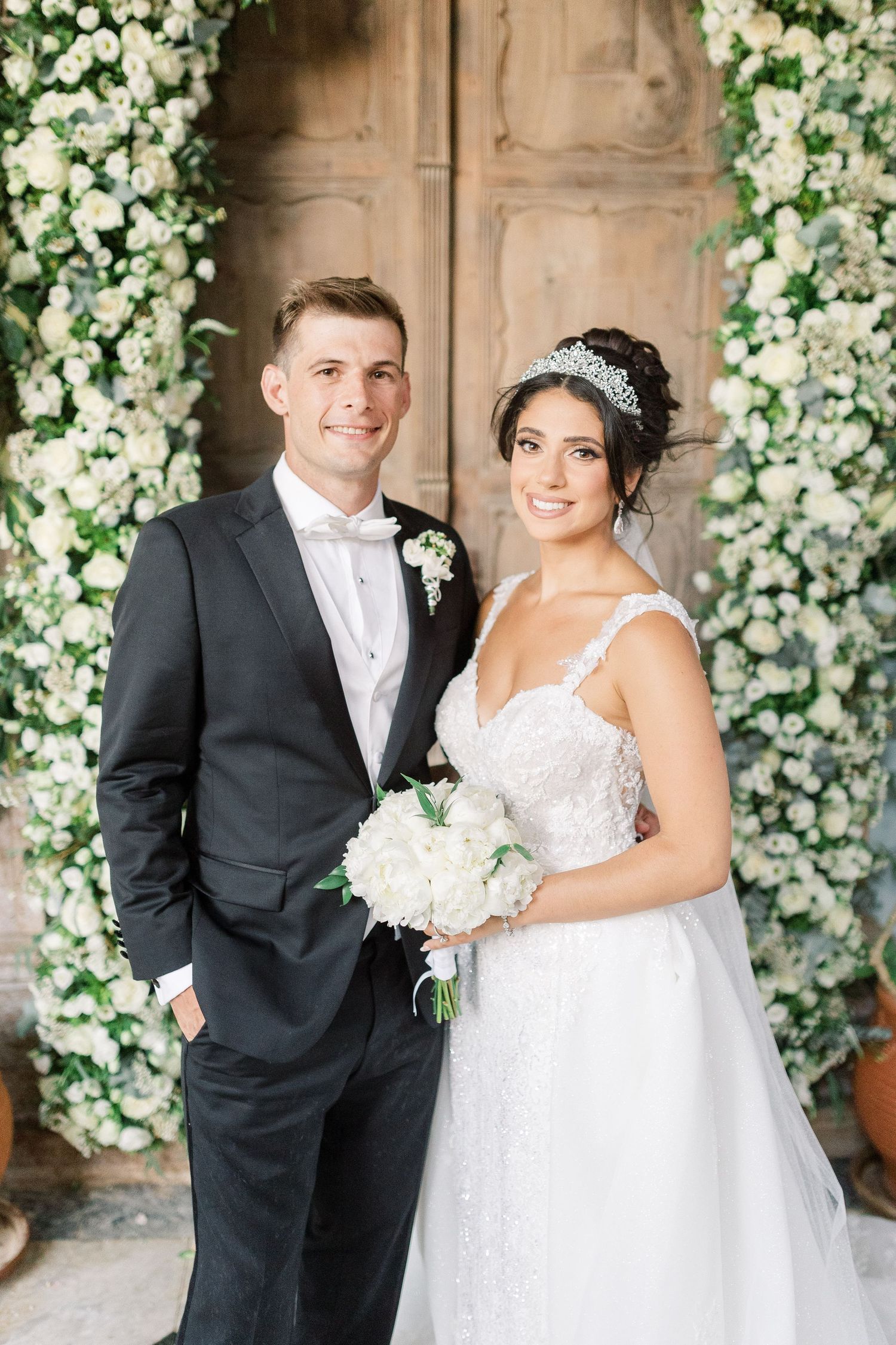 Wedding couple standing against floral arch with white roses and greenery.