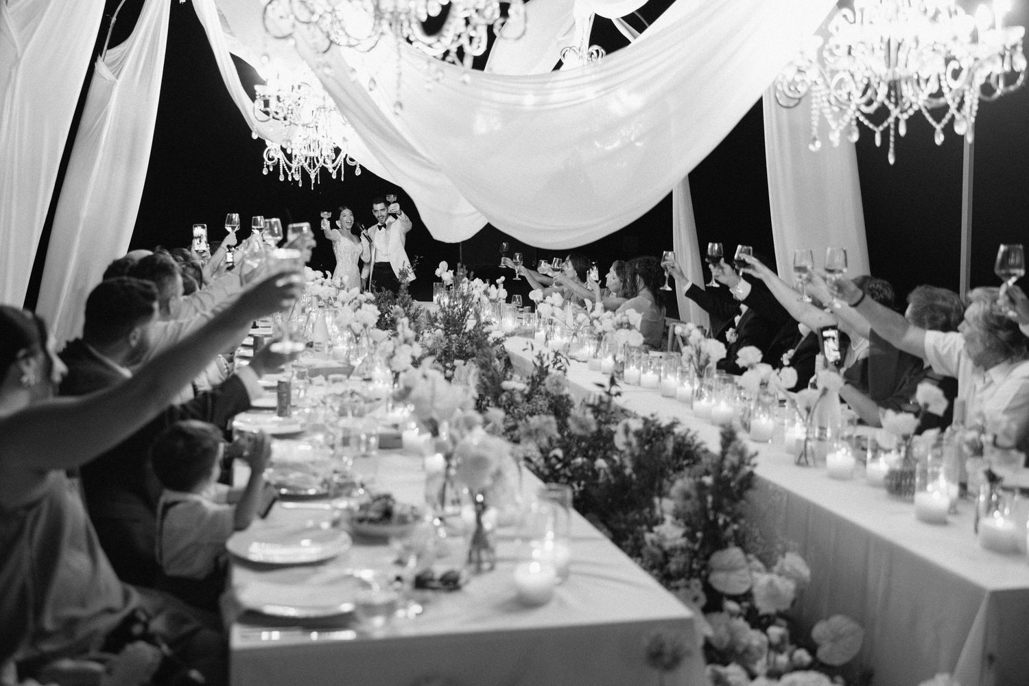 Black and white wedding reception toast with guests raising glasses under elegant chandelier and draped ceiling decor.