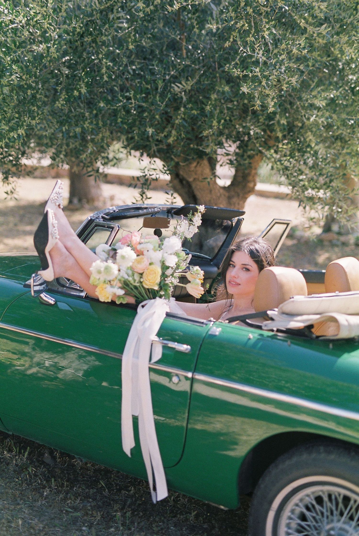 Bride relaxing in a vintage green convertible car adorned with a floral bouquet and white ribbons outdoors.