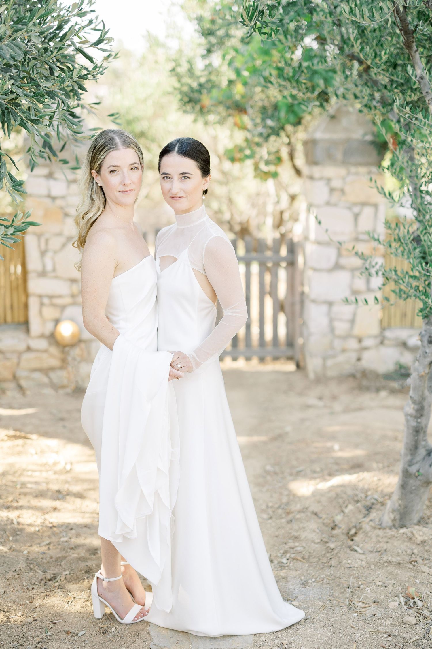Two figures in elegant white dresses pose together against a rustic stone wall and garden backdrop.