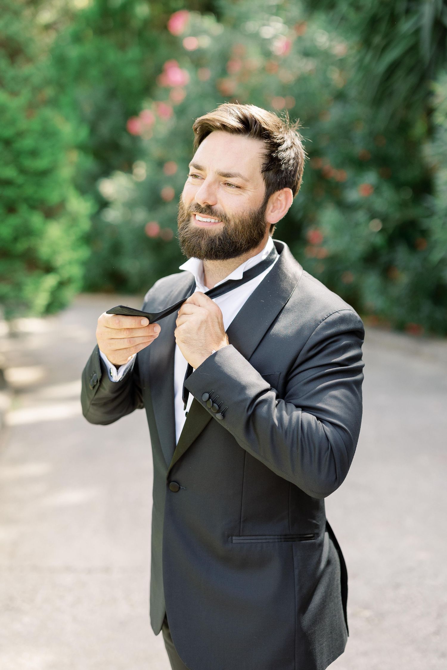 Person in black tuxedo adjusting bowtie while smiling in garden setting.