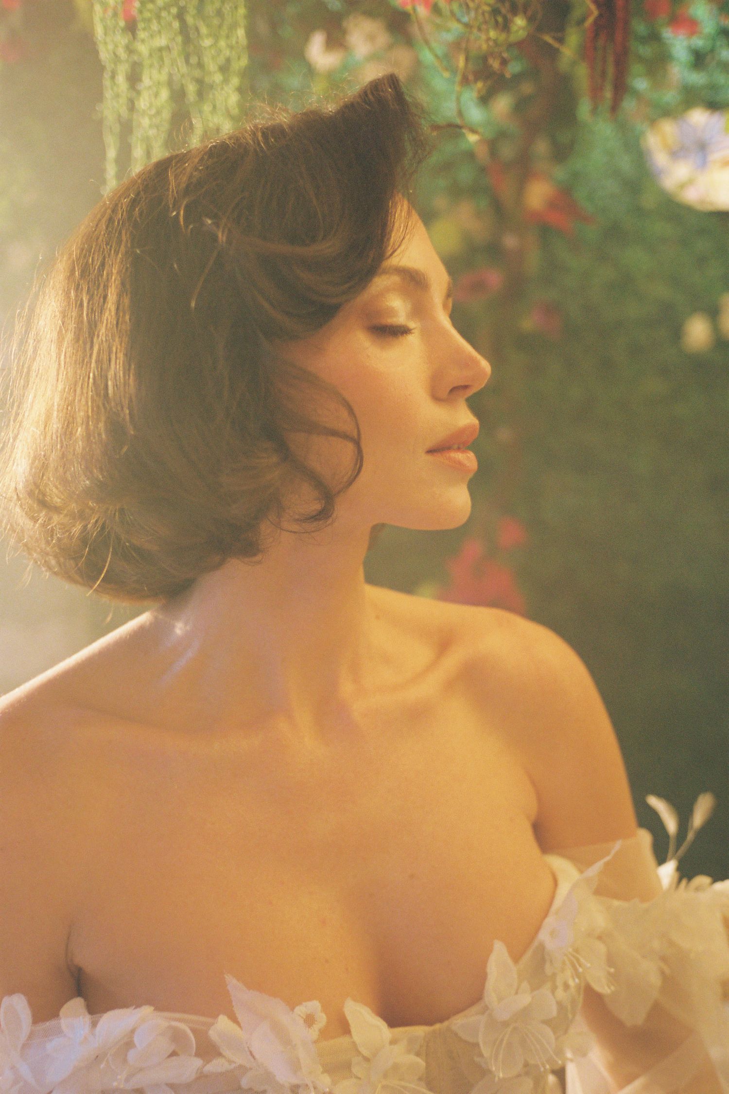 Woman in elegant off-shoulder white dress with flowers, vintage-style portrait with soft focus garden background.