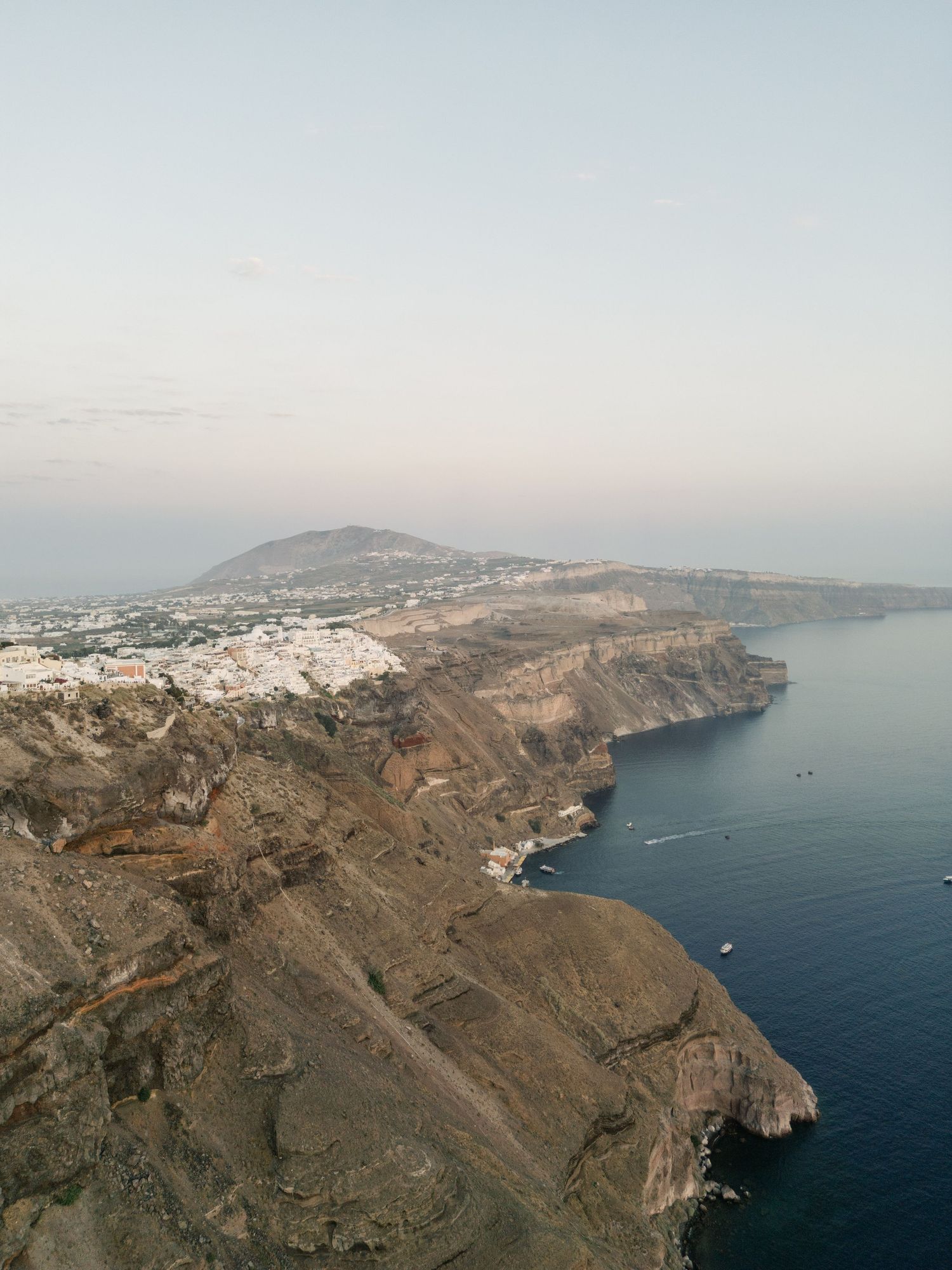 Panoramic aerial view of Santorini's dramatic coastline with white buildings perched on volcanic cliffs above the Aegean Sea.