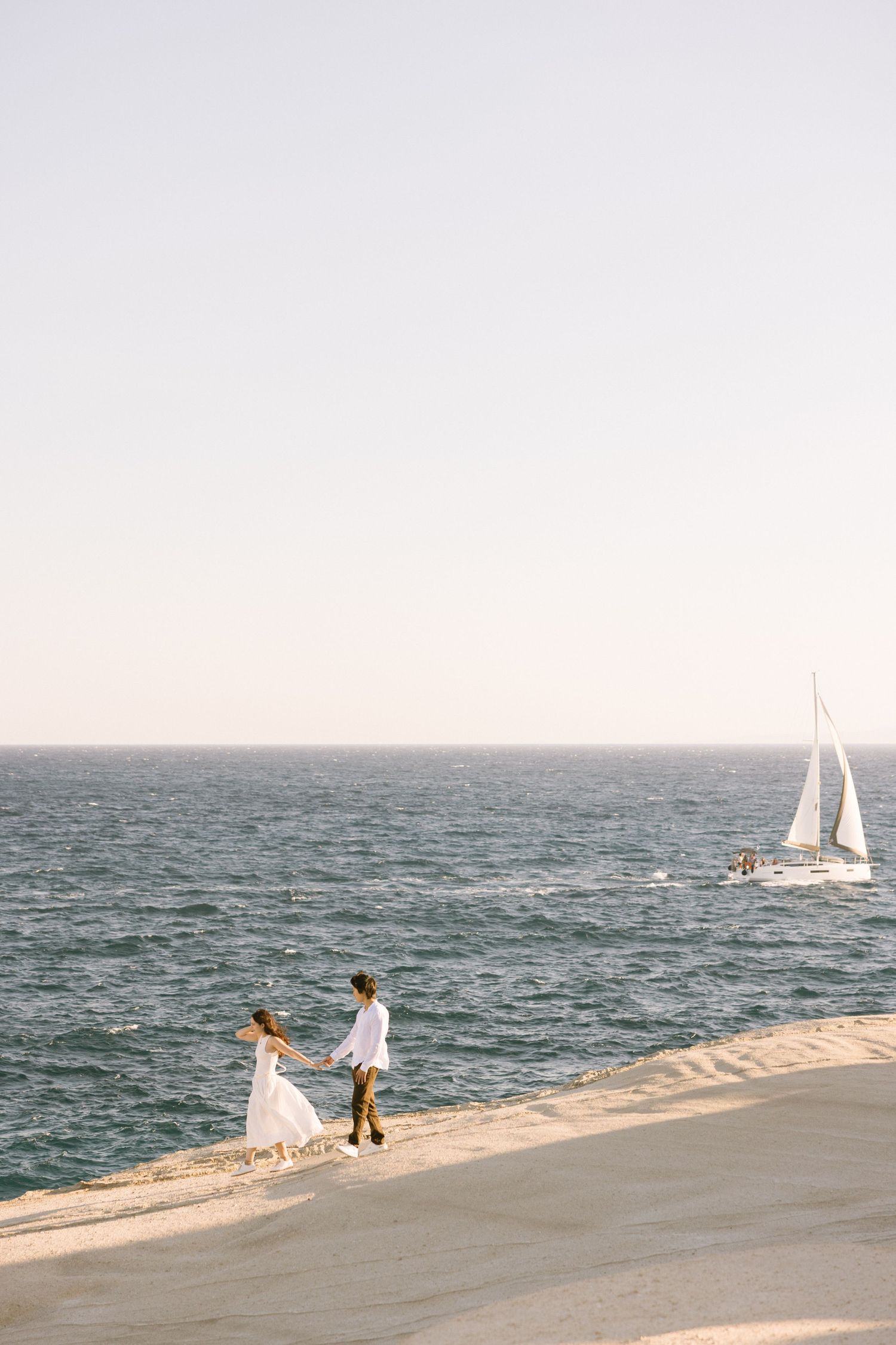 Couple walking along a serene beach shoreline at sunset with sailboats visible in the ocean.