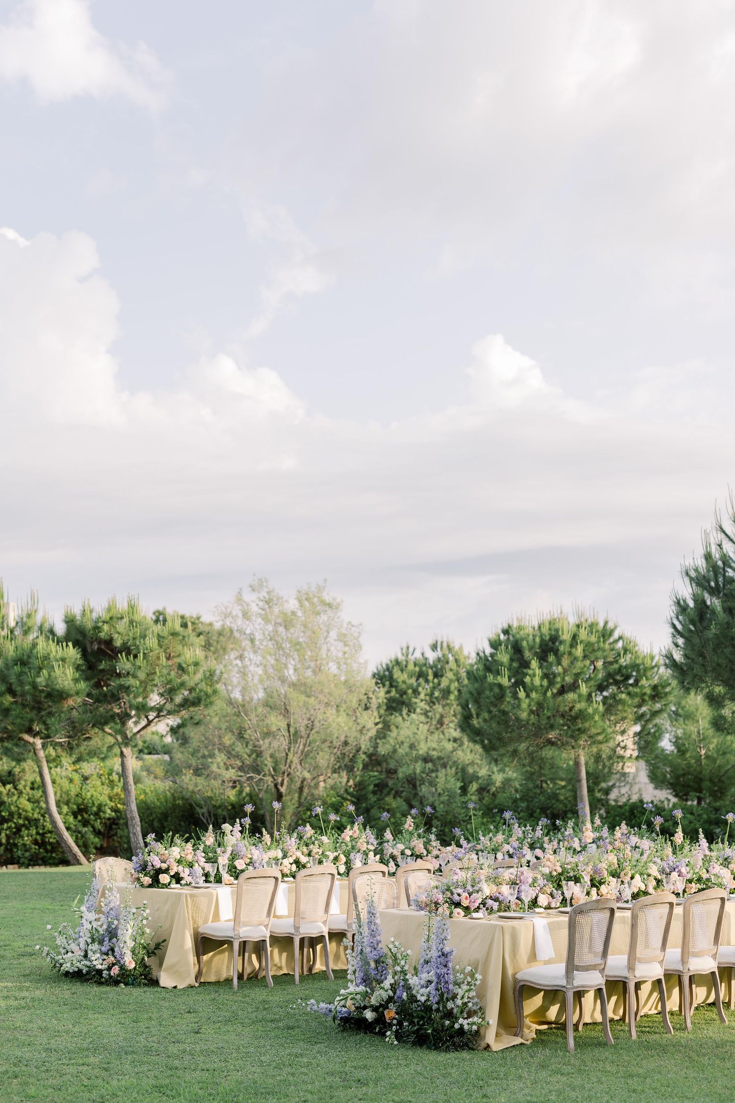 Elegant outdoor wedding reception setup with long tables and chairs arranged on a lush green lawn under blue skies.