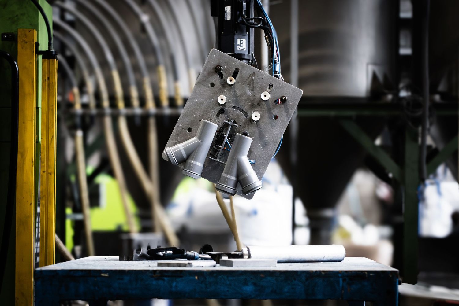 Robotic arm moving small grey HDPE pipes in a manufacturing facility against a busy soft-focus factory background.