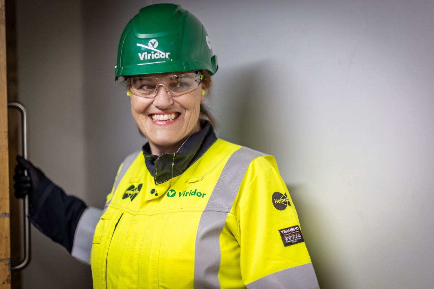 Two workers wearing high visibility yellow vests and green hard hats stand in a warehouse setting.