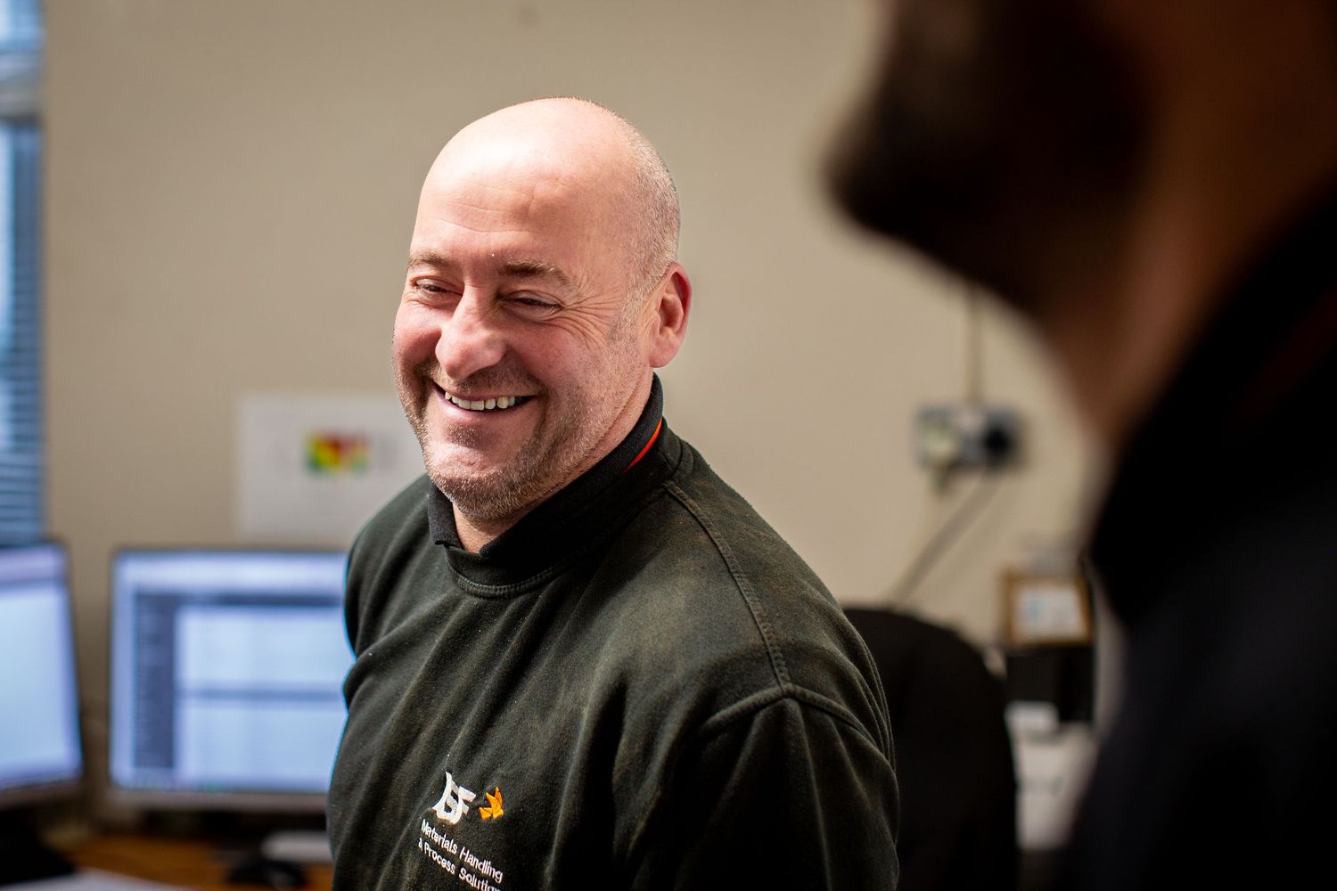 A person in a black shirt smiles while working at a desk in an office environment.
