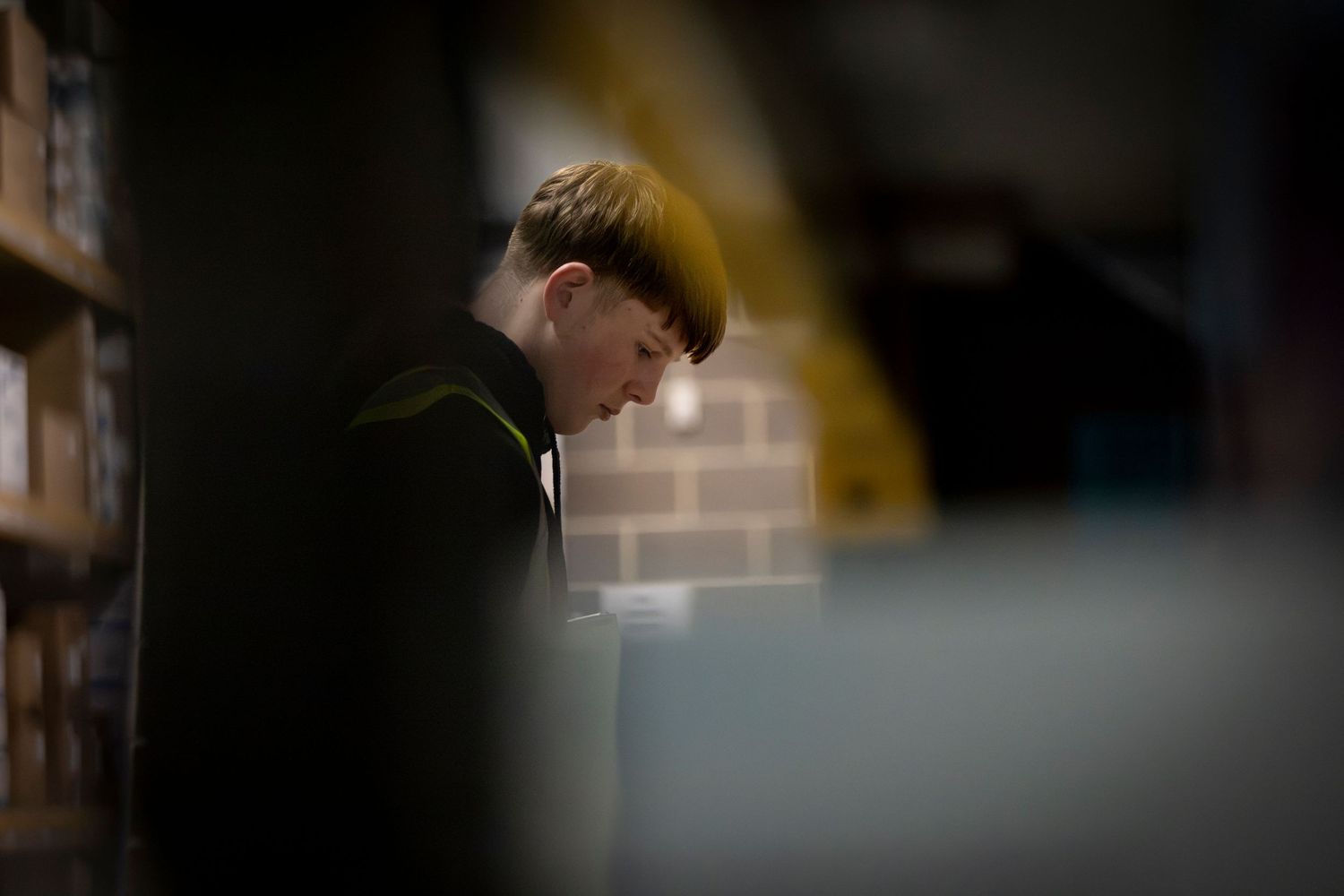 Young man reading intently in a dimly lit library, surrounded by bookshelves and brick walls.