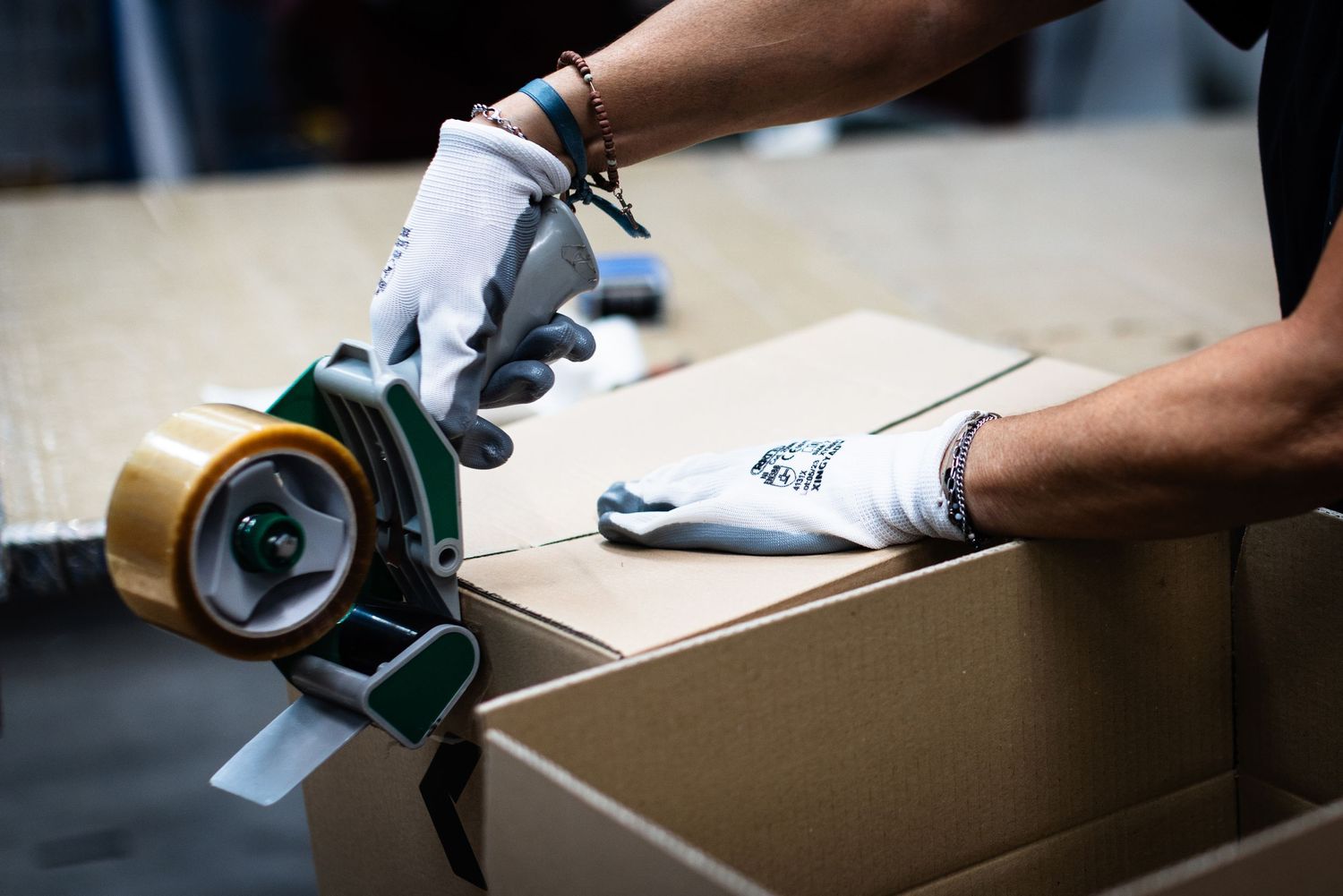 Worker in gloves sealing a cardboard box with packing tape dispenser in a warehouse.