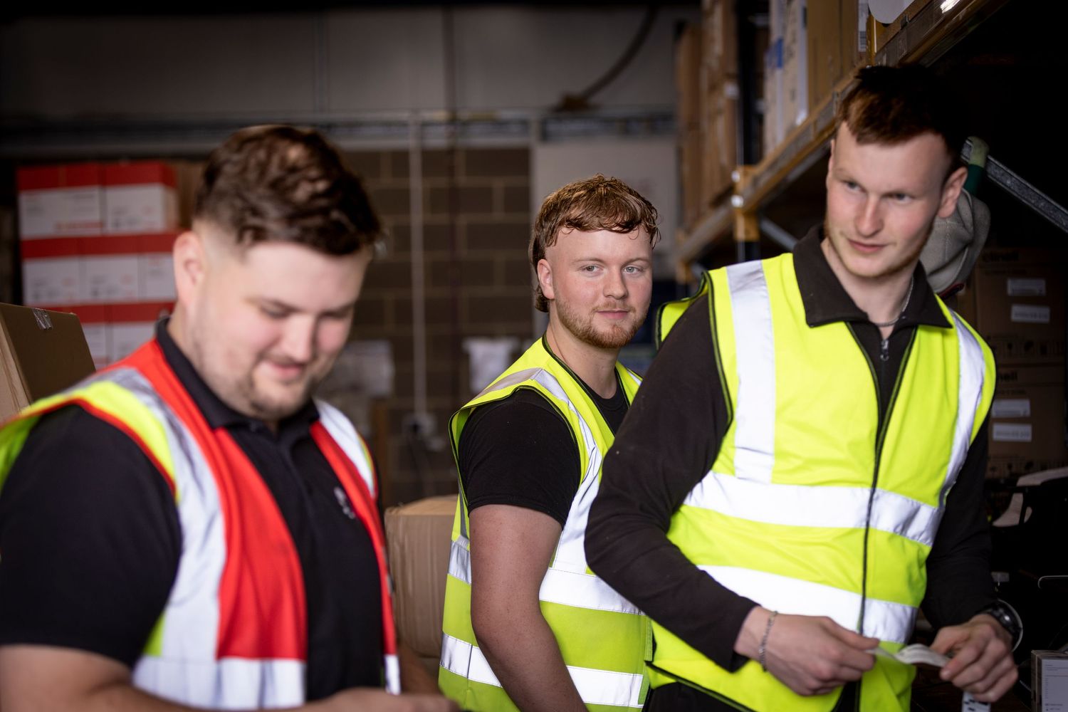 Three warehouse workers in high-visibility vests reviewing documents in a storage facility.