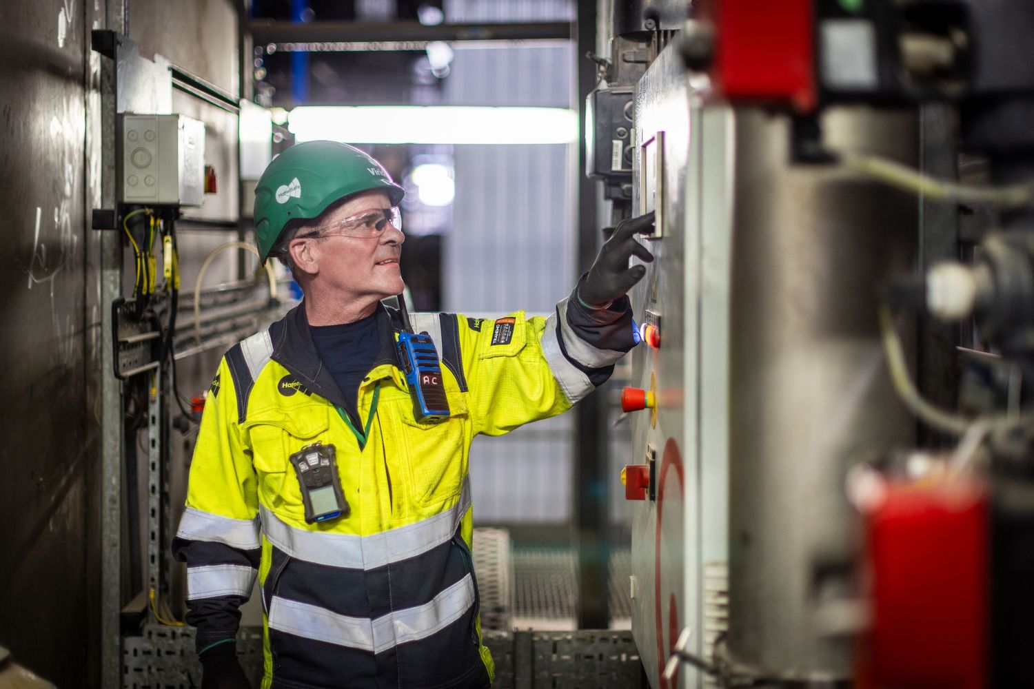 Industrial worker in safety gear and green cap inspecting equipment in a manufacturing facility.