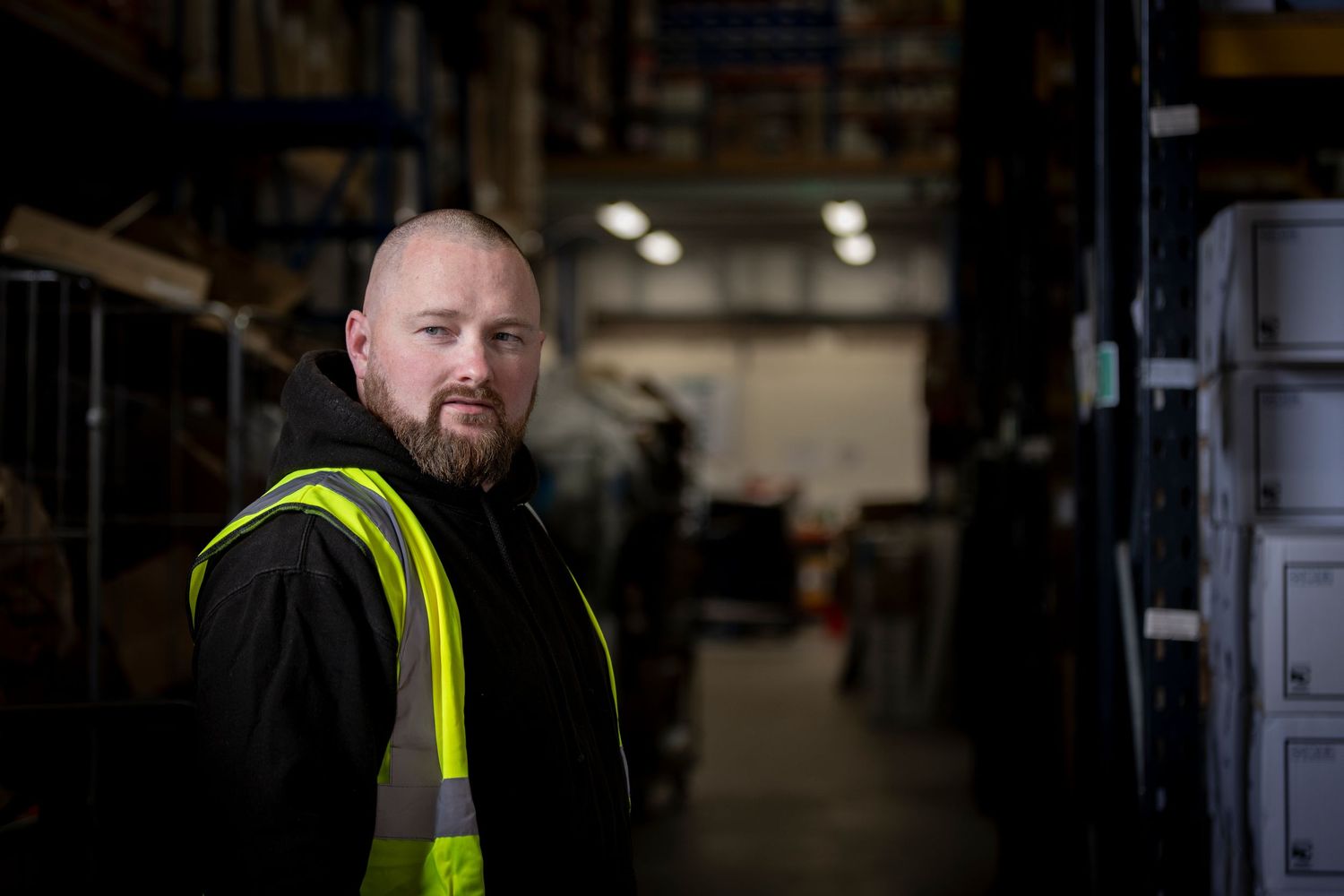 Warehouse worker in high-visibility vest stands confidently among industrial shelving in a large storage facility.