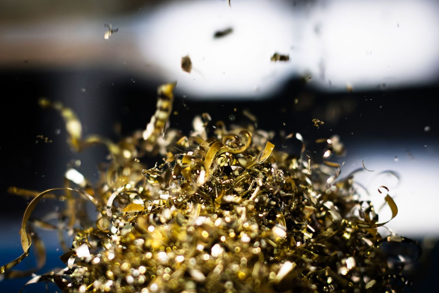 Metallic brass shards fall like confetti into a sparkling pile on dark background with dramatic back lighting..