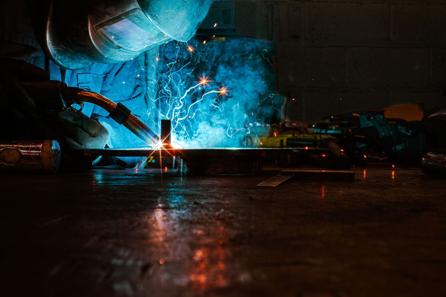 Blue welding sparks illuminate a dark industrial workspace with metal equipment and machinery in the background.