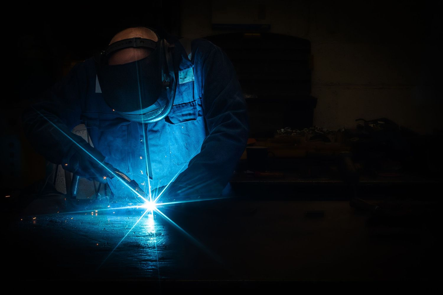 A welder works in darkness with bright blue welding sparks illuminating the metalwork in dramatic lighting.