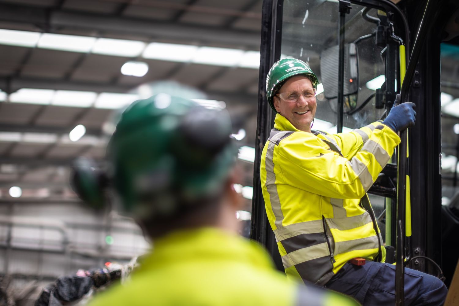Workers in safety vests and hard hats operate machinery in an industrial warehouse setting.