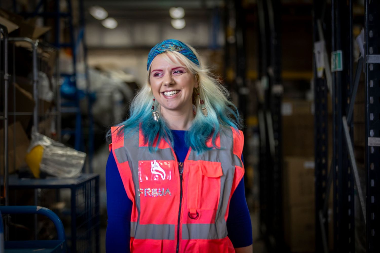 Woman with blue hair wearing a red hi-vis vest smiles in a warehouse storage facility.