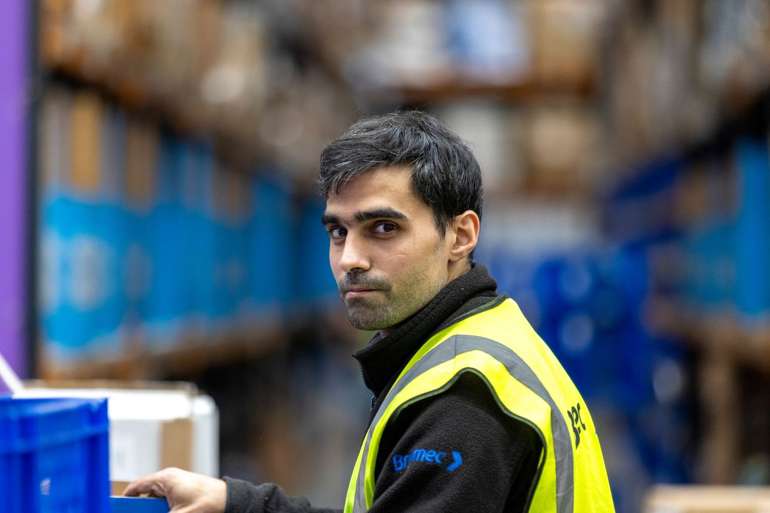 Warehouse worker in yellow safety vest stands among shelving racks in a large distribution center.