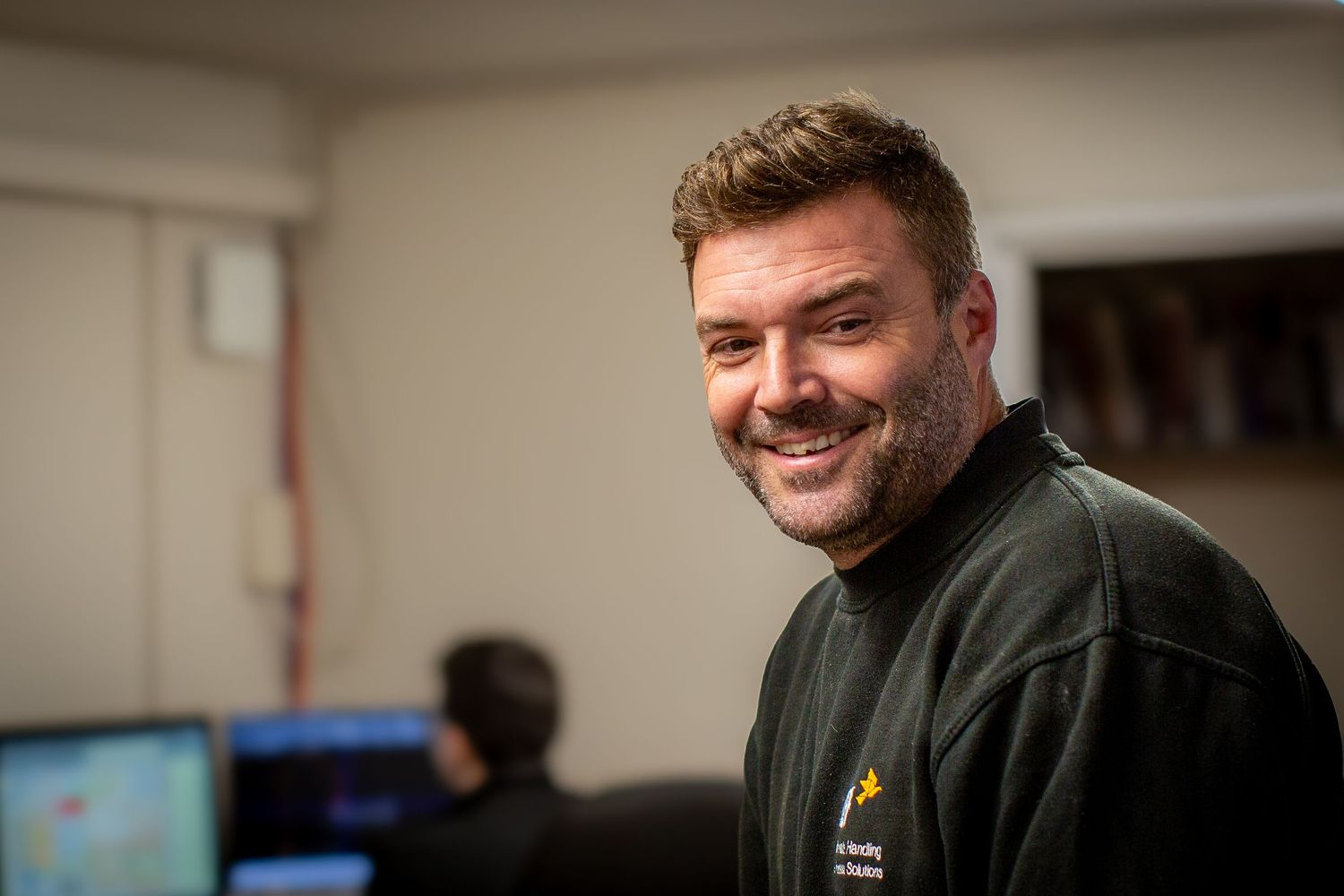 A smiling person in an olive green jacket sits in an office environment with computer monitors visible in the background.