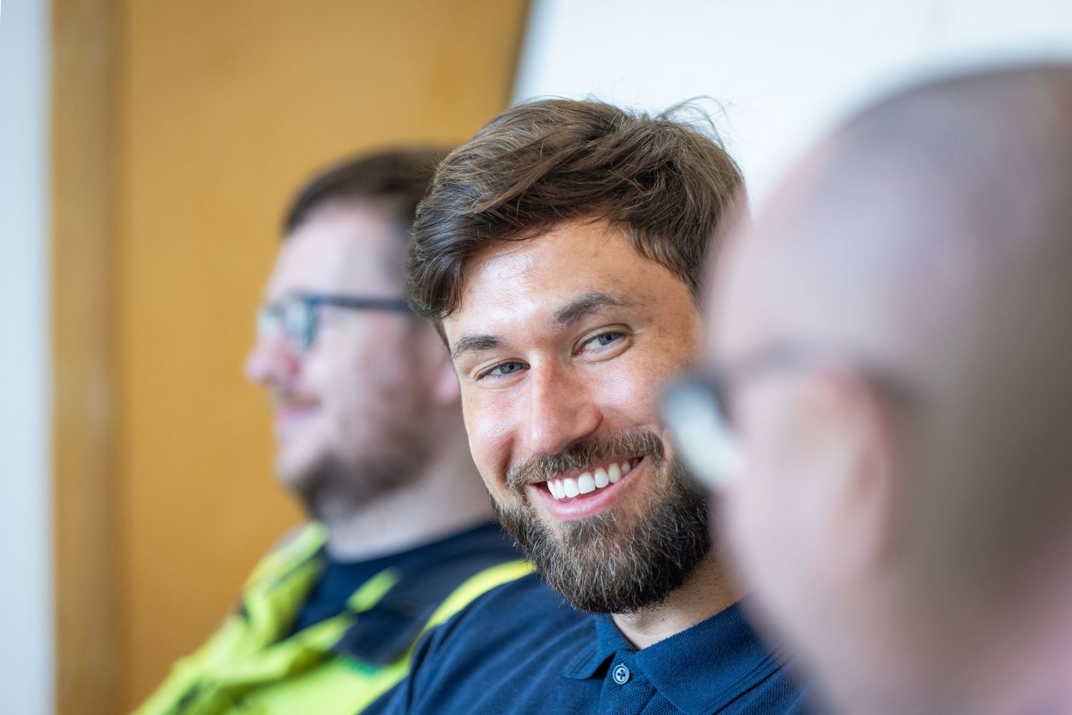 A person with a beard smiles warmly while sitting with others in a casual indoor setting.