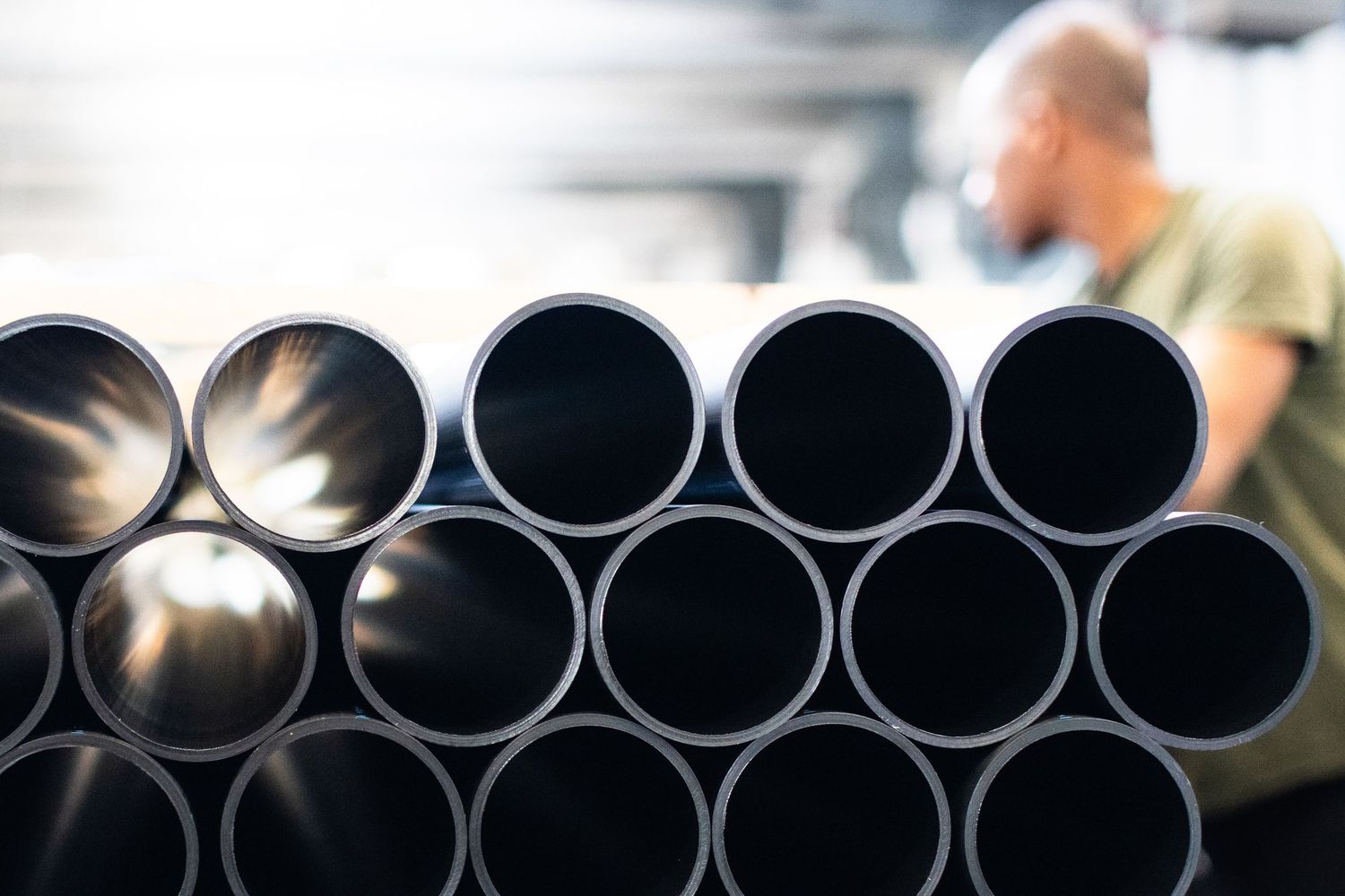 Rows of large black industrial HDPE pipes stacked in parallel formation with softly focused worker in motion behind.