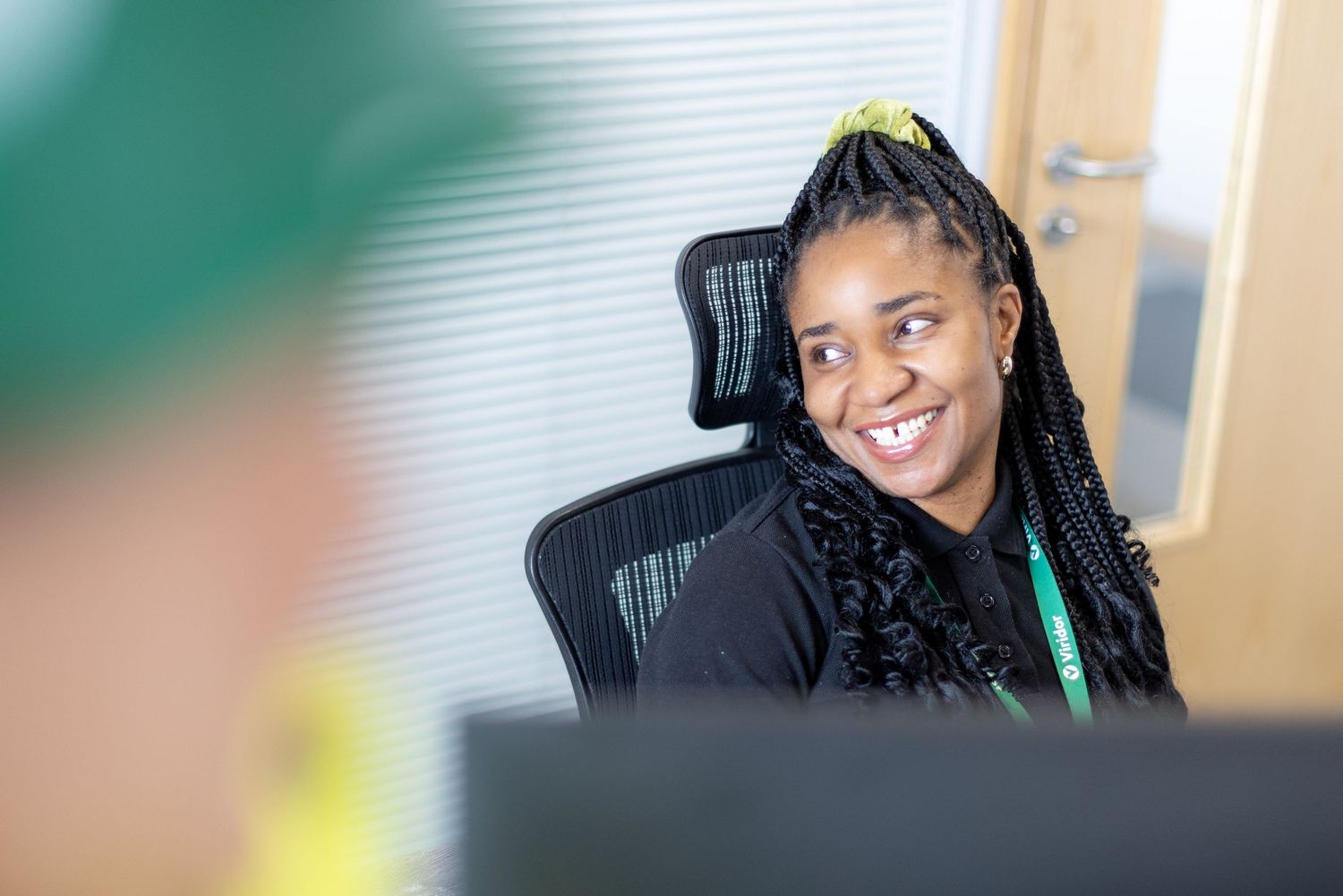A smiling professional sits at a desk in an office, wearing a black top with braided hair and a warm expression.