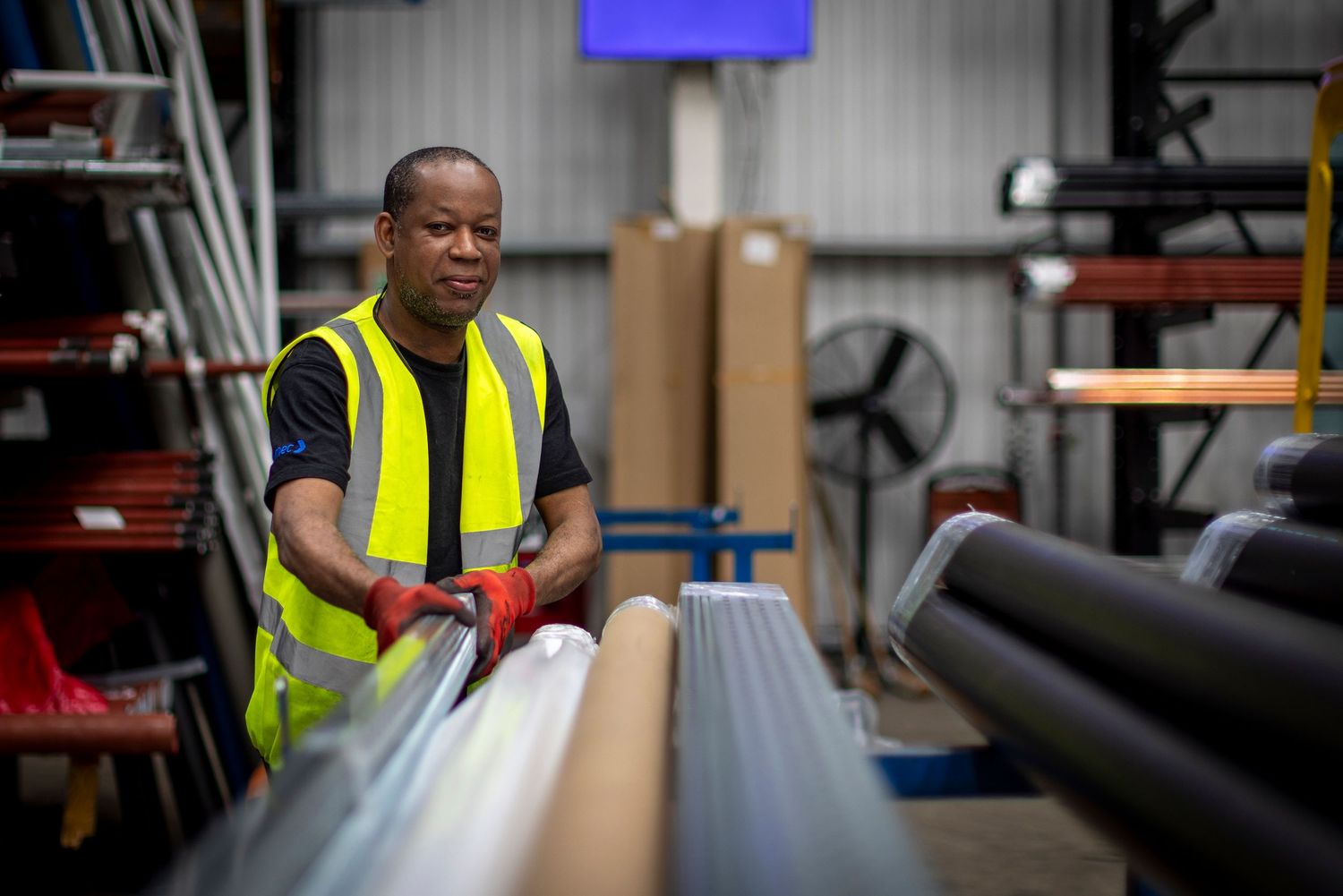 Worker in yellow safety vest organizing pipes and materials in an industrial warehouse storage facility.
