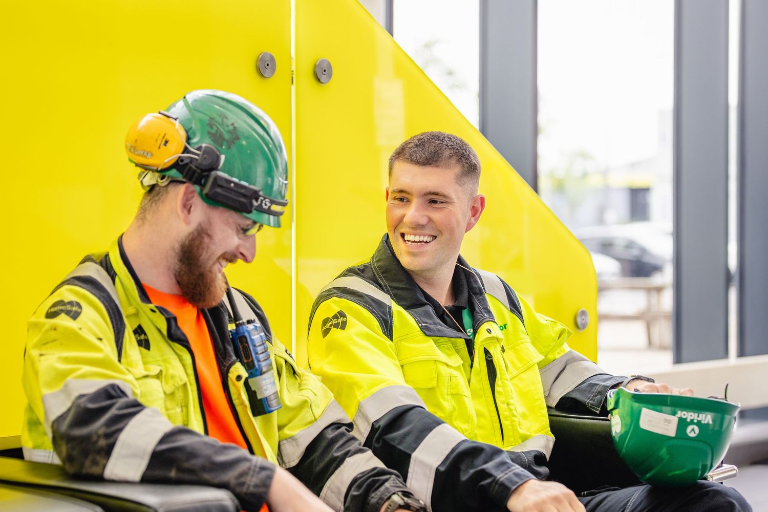 Two workers in high visibility yellow safety gear and hard hats share a laugh while taking a break on the job.