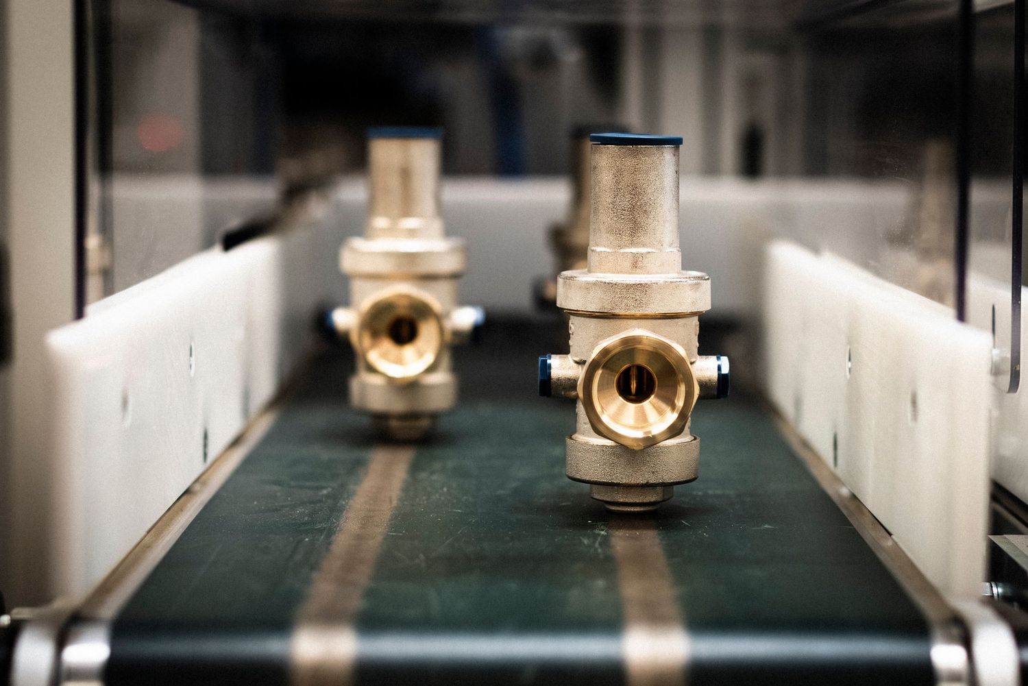 Two brass valve housings moving towards the viewer along a green conveyor belt in a dark moody lighting.