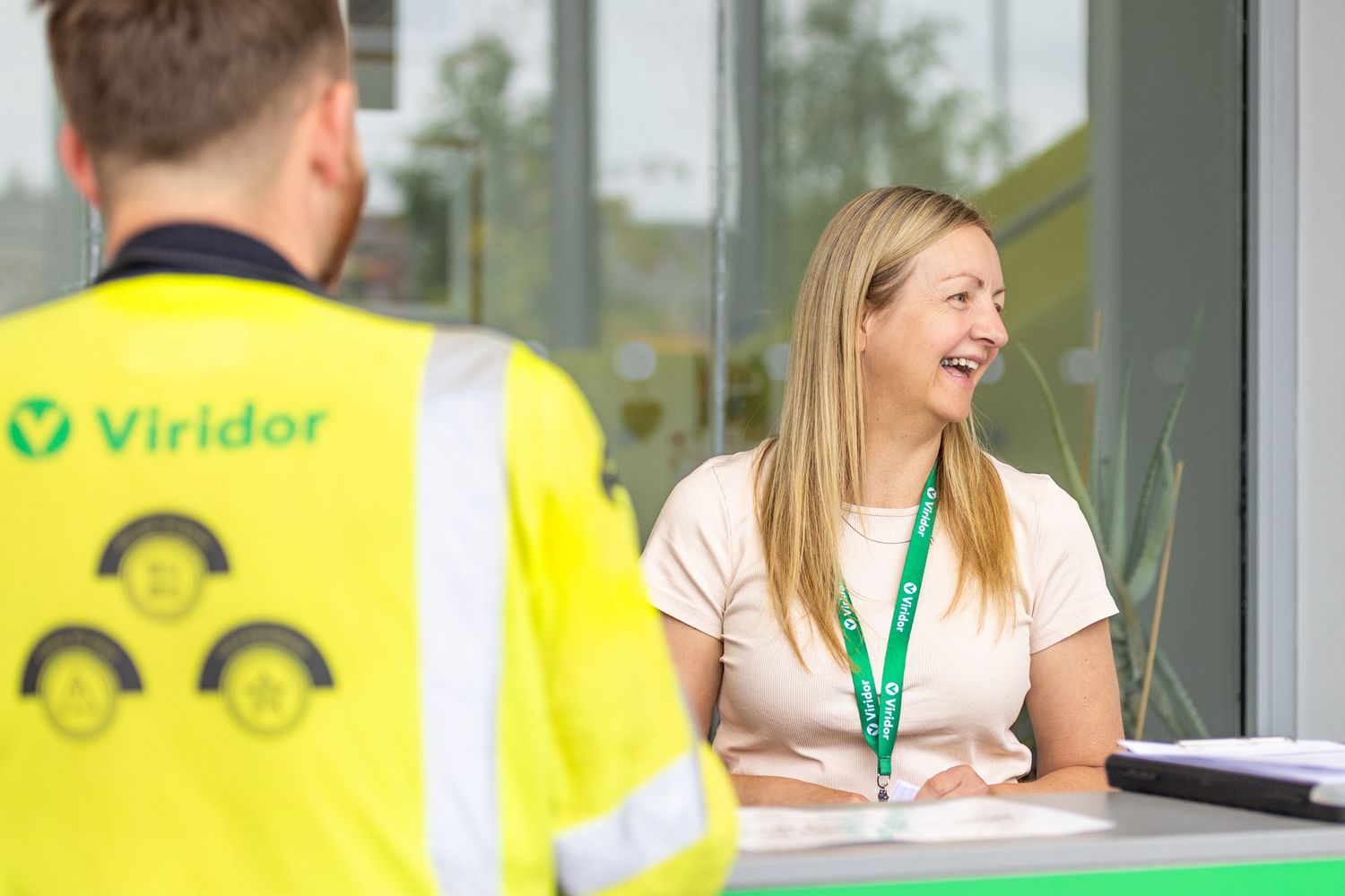 A customer service interaction at a desk between someone in a yellow Viridor vest and a staff member wearing a lanyard.