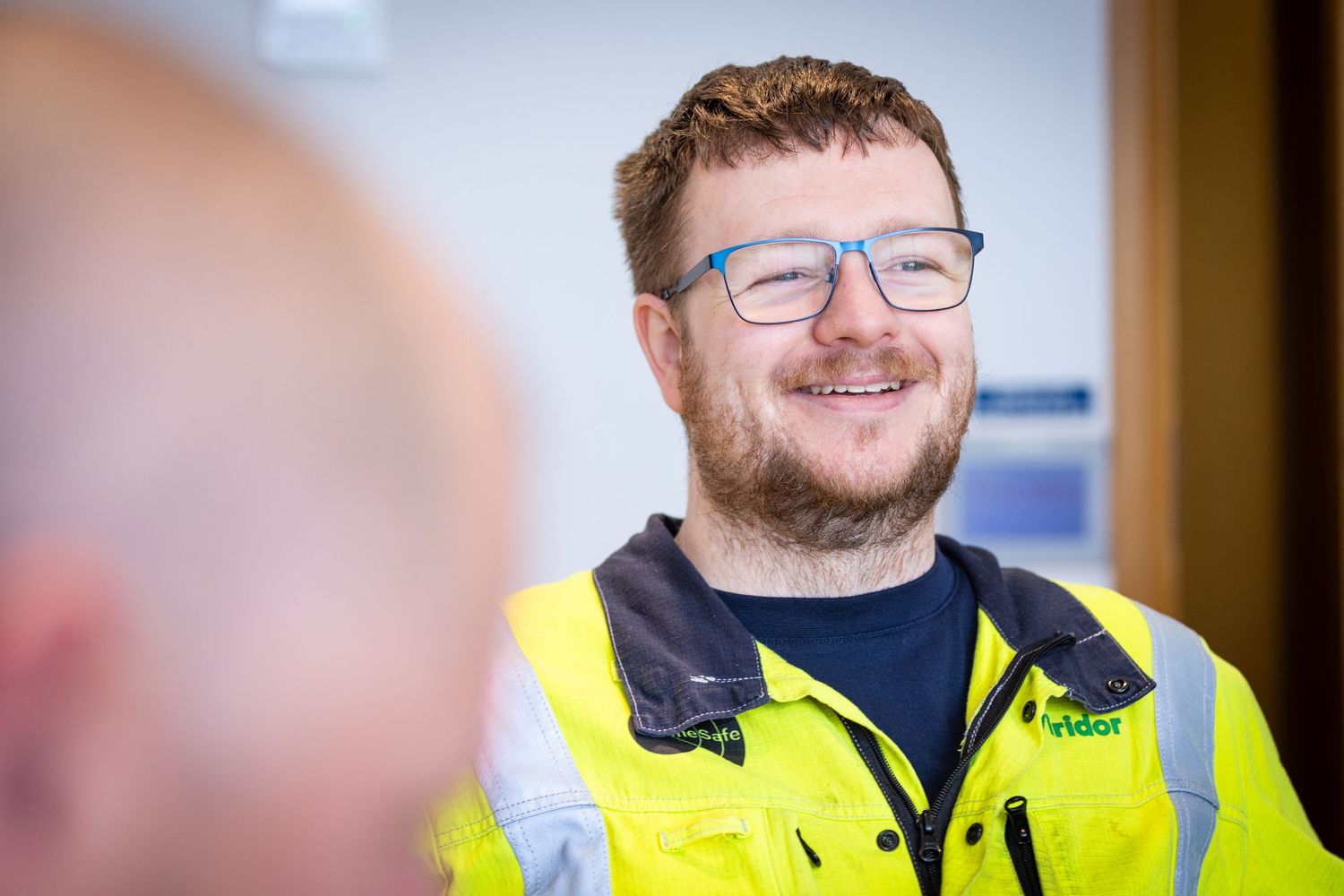 Construction worker wearing high visibility vest and glasses smiles while standing in indoor workspace.