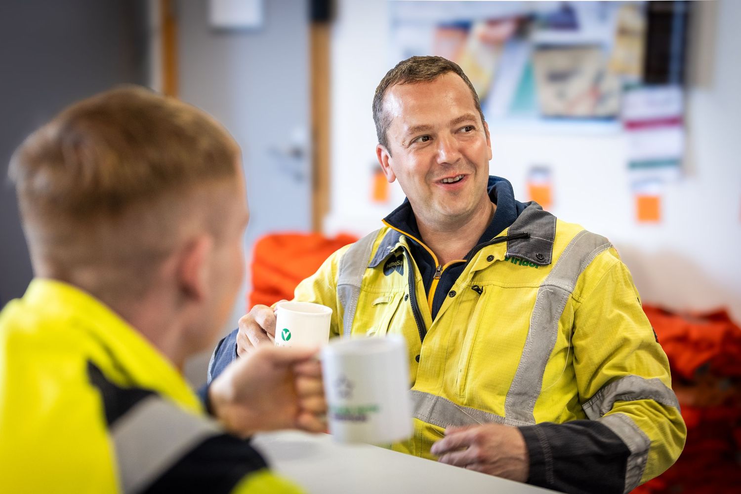 Workers in high visibility yellow jackets gather around a table during a break or meeting at a workplace.