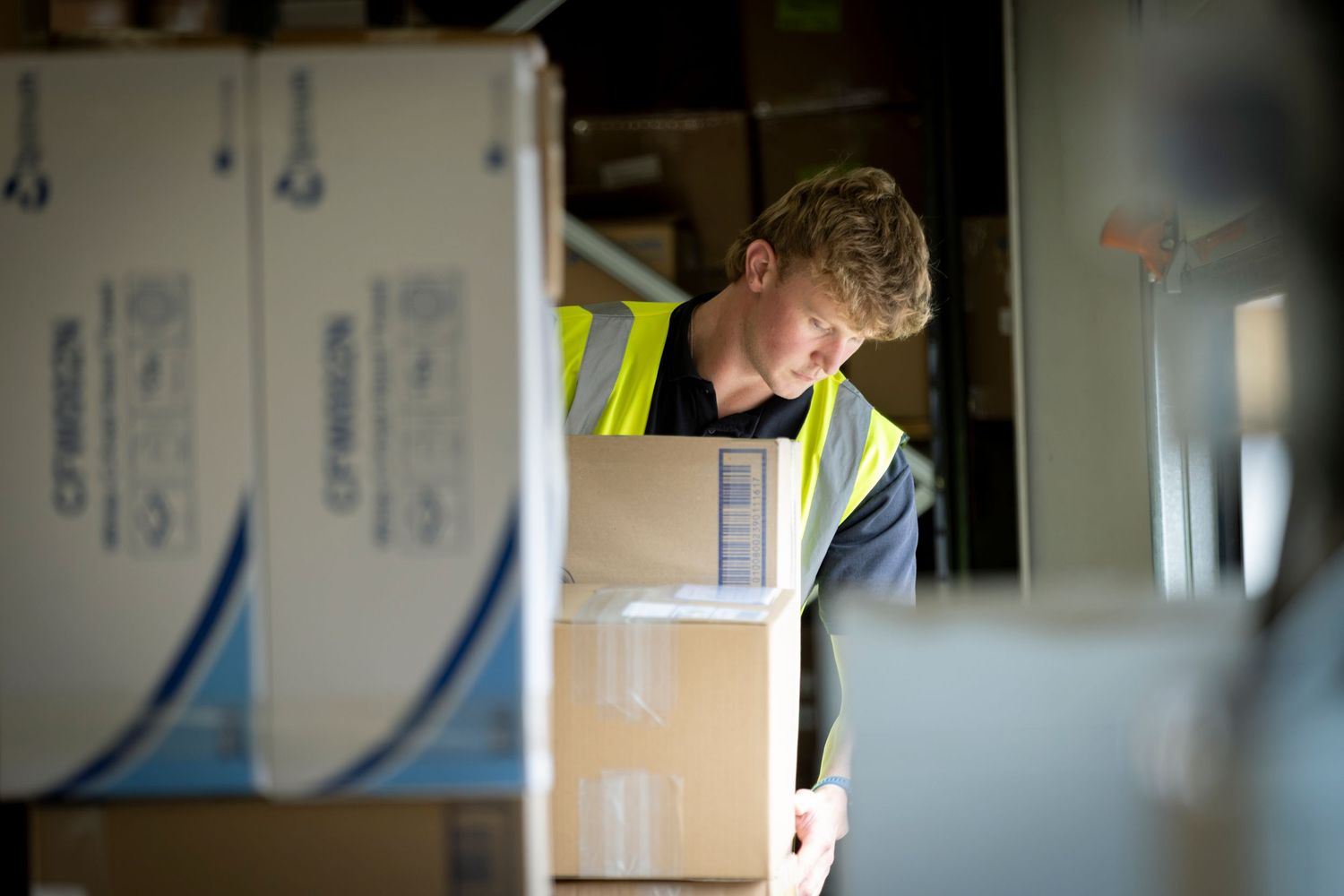 Warehouse worker in yellow safety vest unloading cardboard boxes in a busy storage facility.