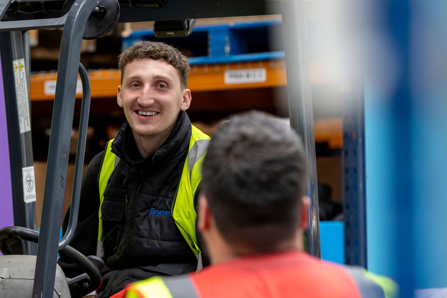 Smiling warehouse worker in yellow safety vest operating a forklift in a busy distribution center.