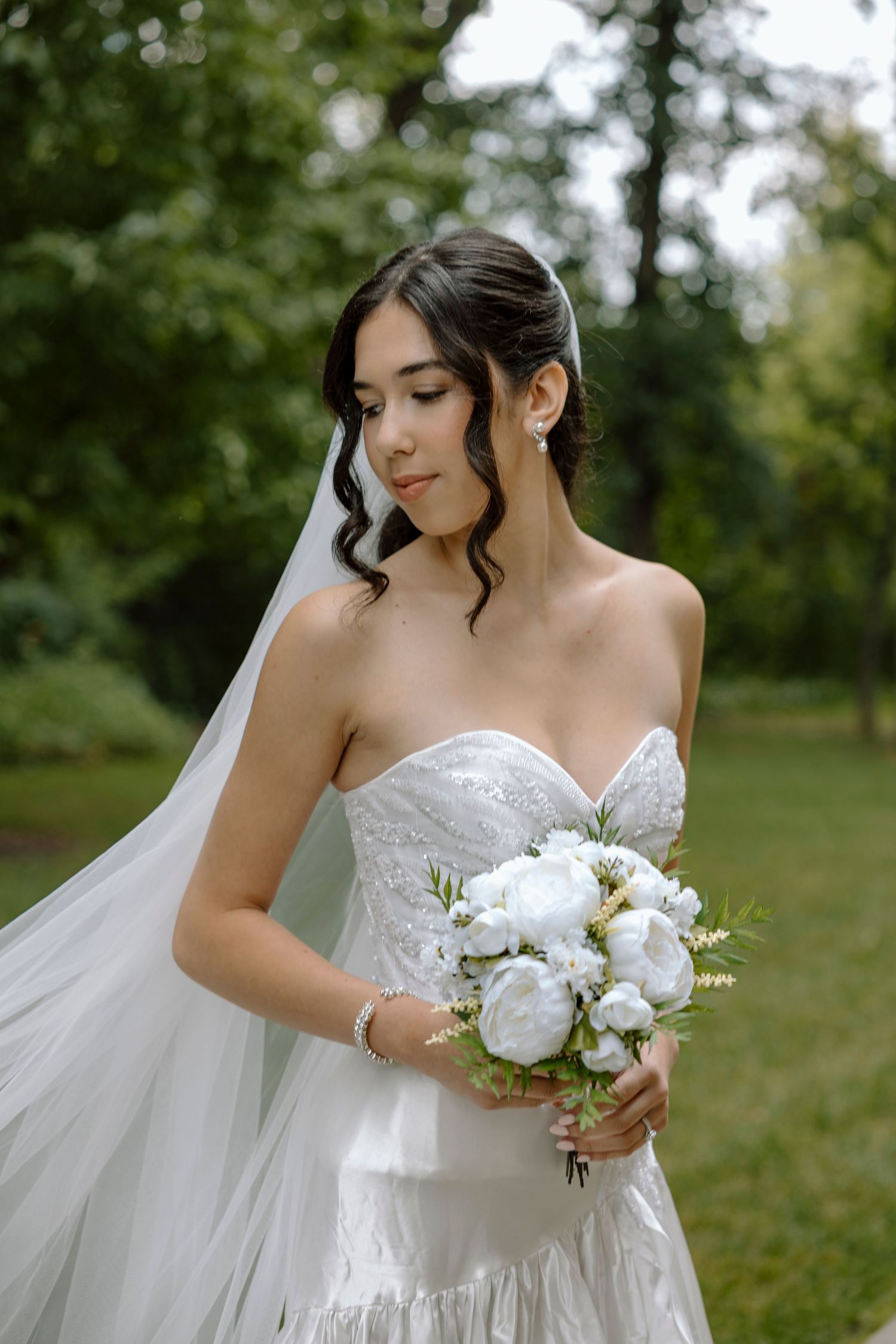 Elegant bride in strapless white lace gown holding white peony bouquet with flowing veil in lush green garden.
