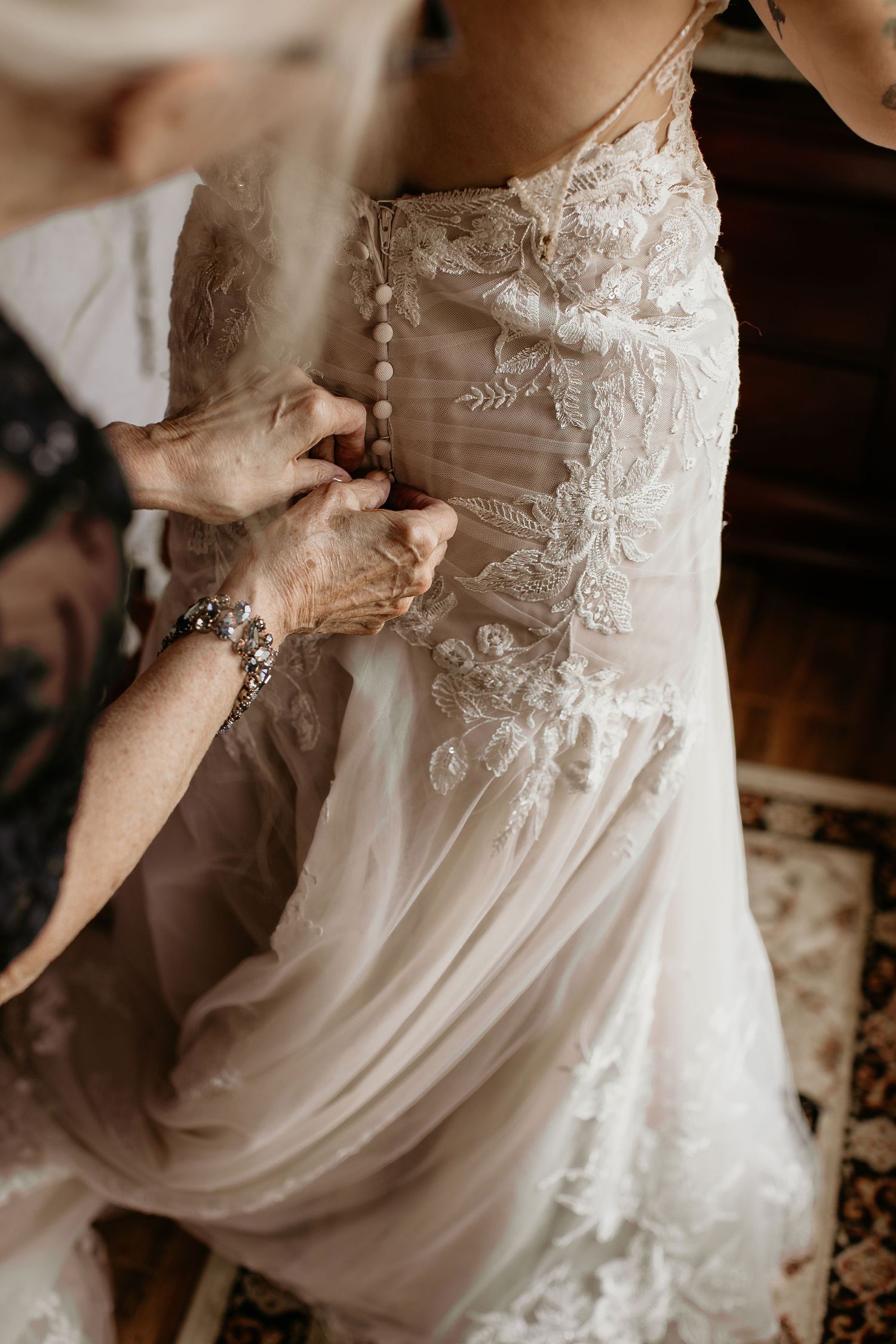 Elderly woman fastening pearl buttons on back of lace wedding dress, bridal preparation moment.